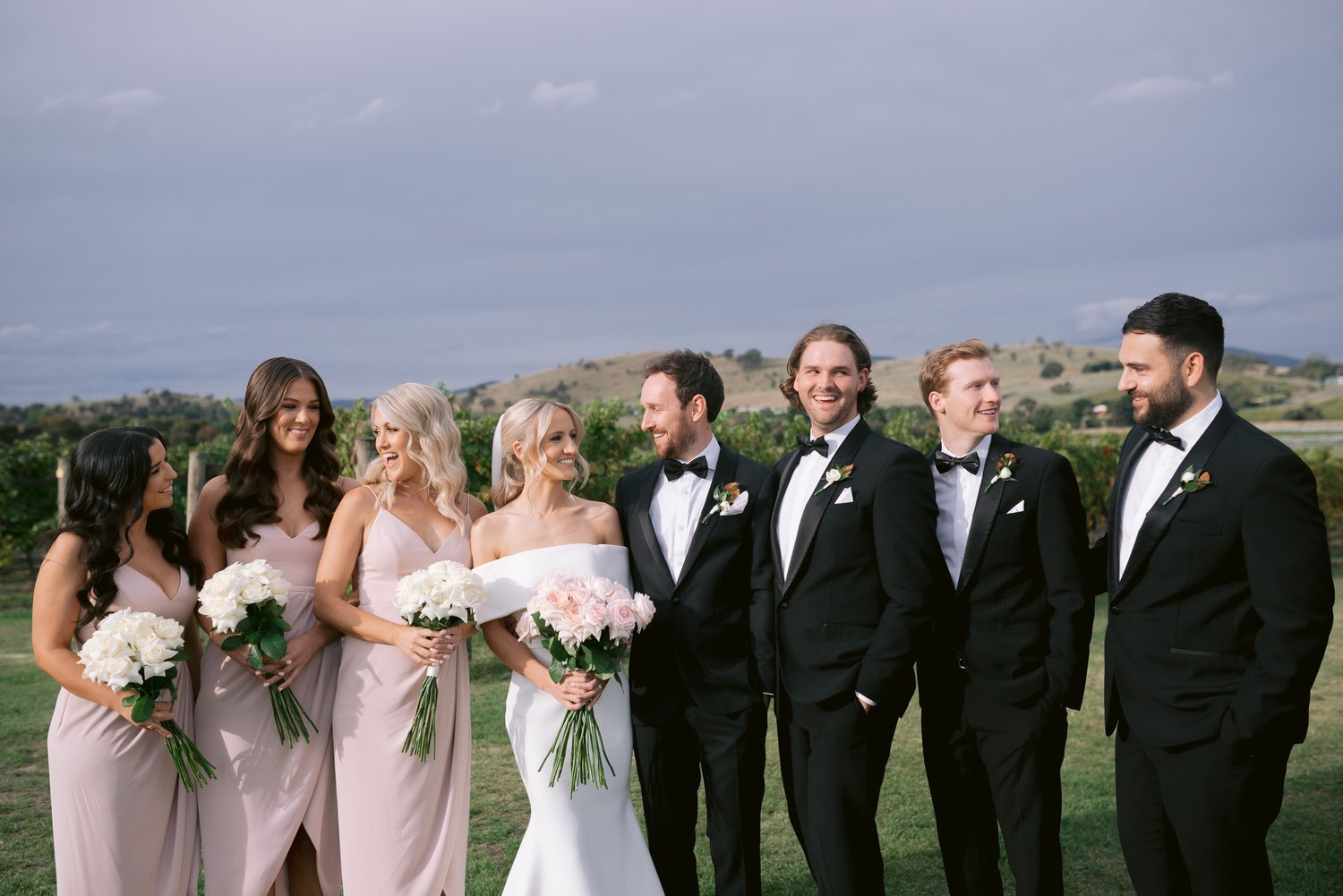 Wedding party group photo outdoors with bridesmaids in pink dresses and groomsmen in black suits.