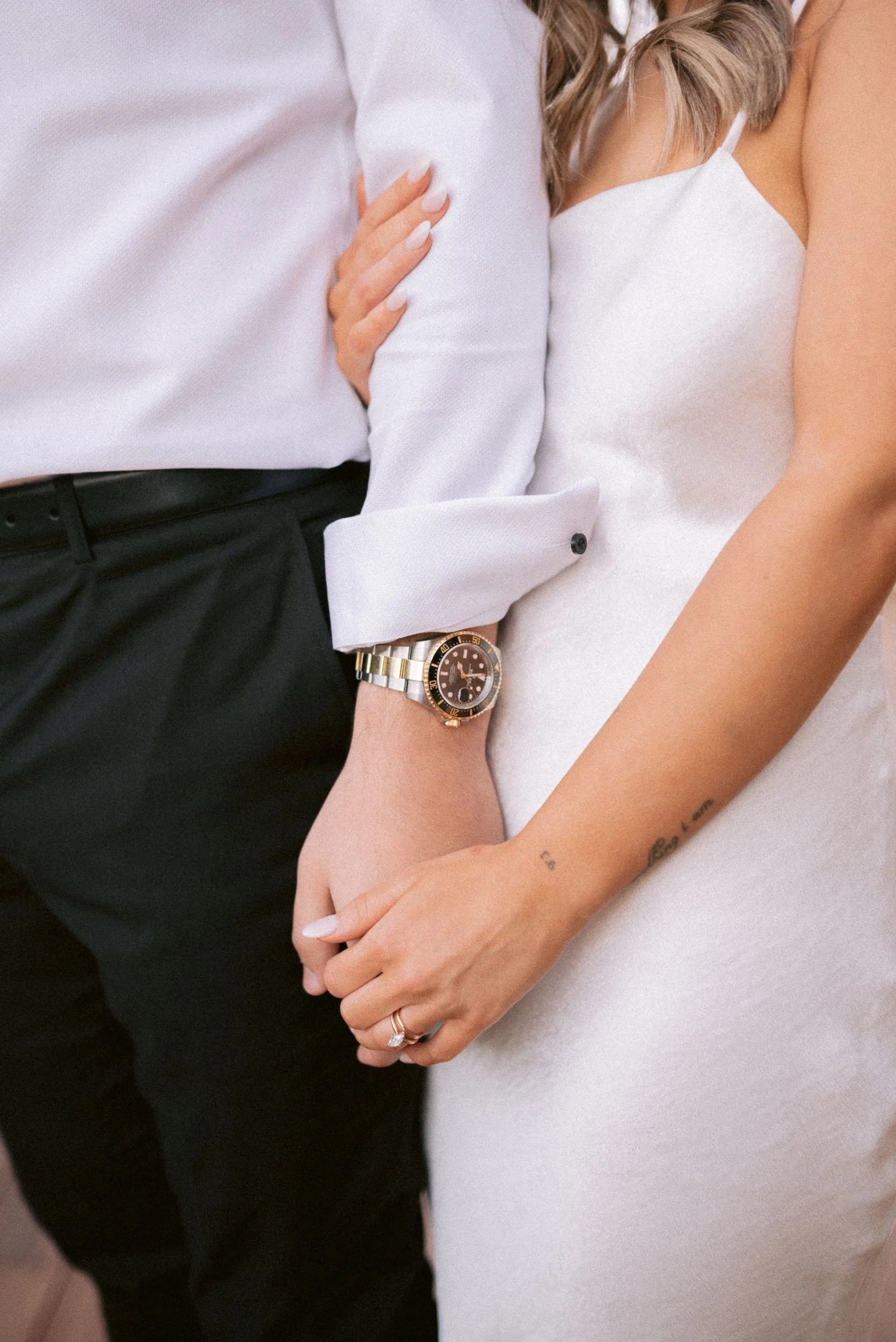 A couple holding hands, with the woman's hand resting on the man's arm, showing a wedding ring and a watch, and a tattoo on her wrist.