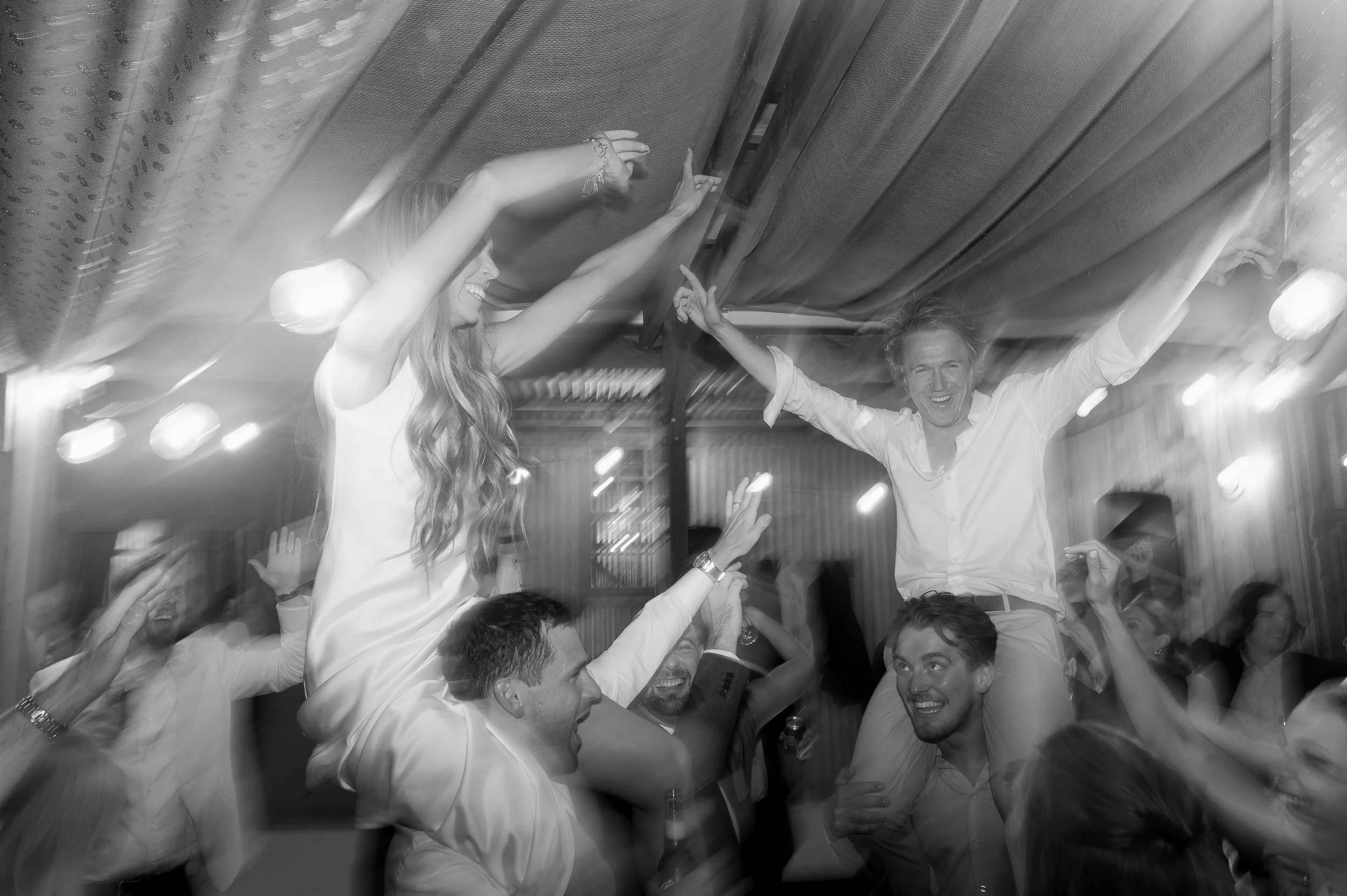 People celebrating and dancing at a party, with two women on others' shoulders, smiling and raising their arms.