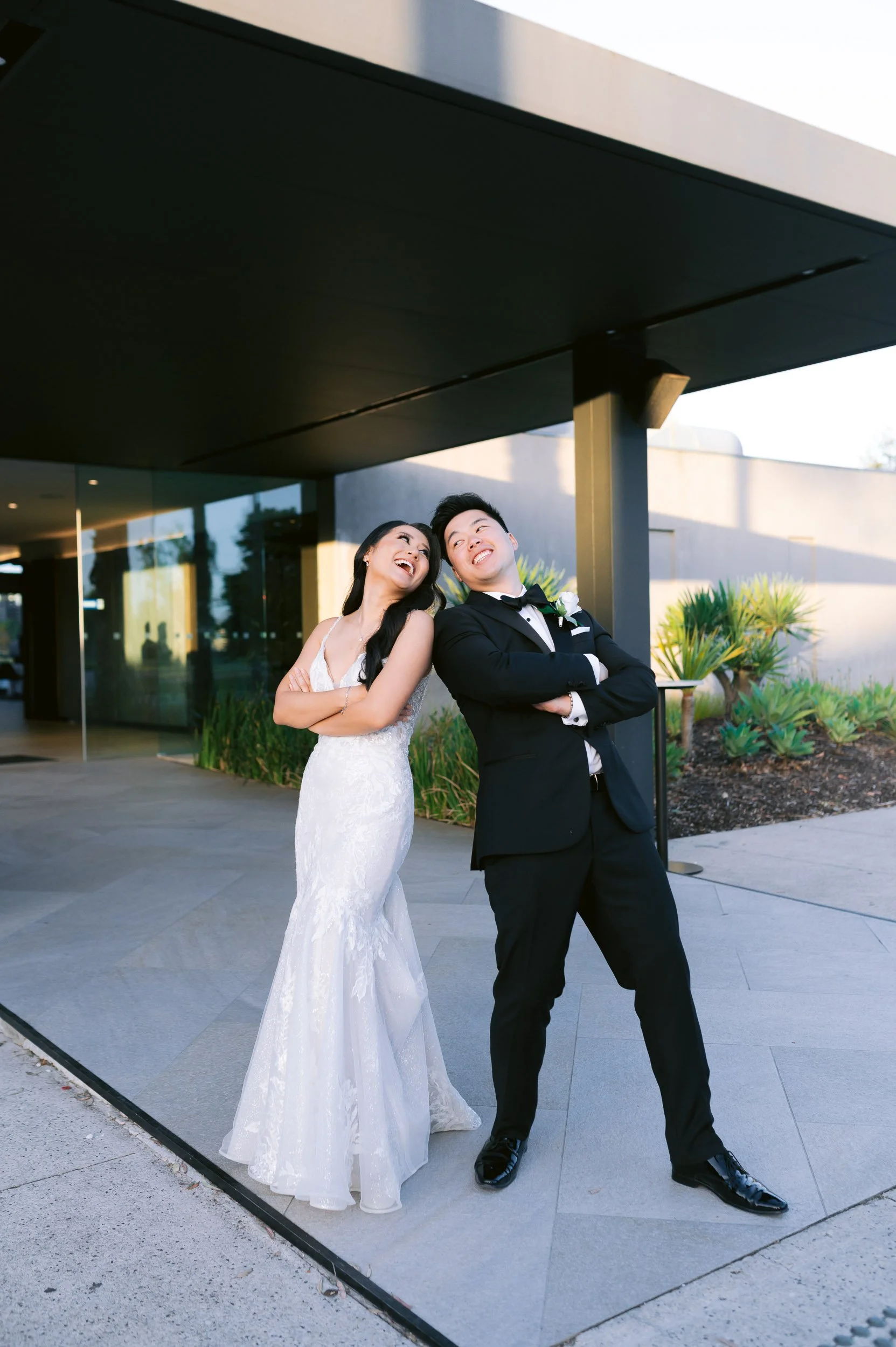 A joyful couple in formal attire posing playfully outside, with the woman in a white wedding dress and the man in a black tuxedo.