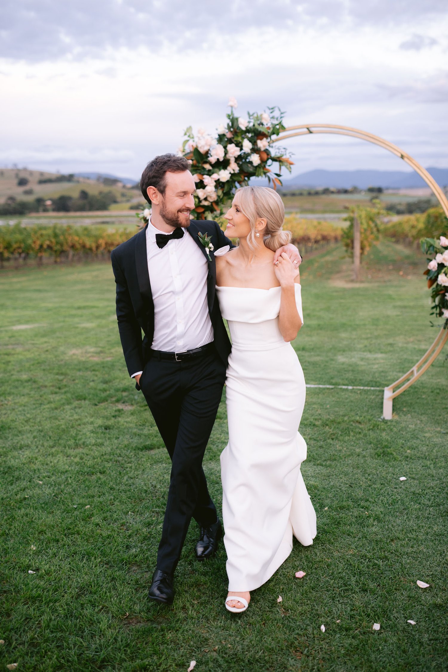 Bride and groom walking on grass near a floral arch, vineyard background.