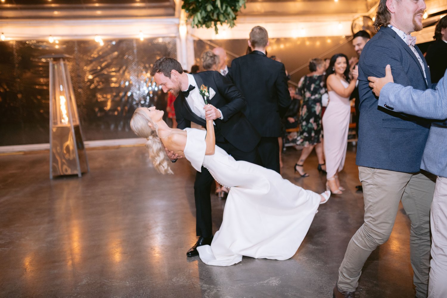 A couple dancing at a wedding reception, with the groom in a suit and the bride in a white dress, surrounded by guests on the dance floor. A heater is visible in the background with decorative lights.