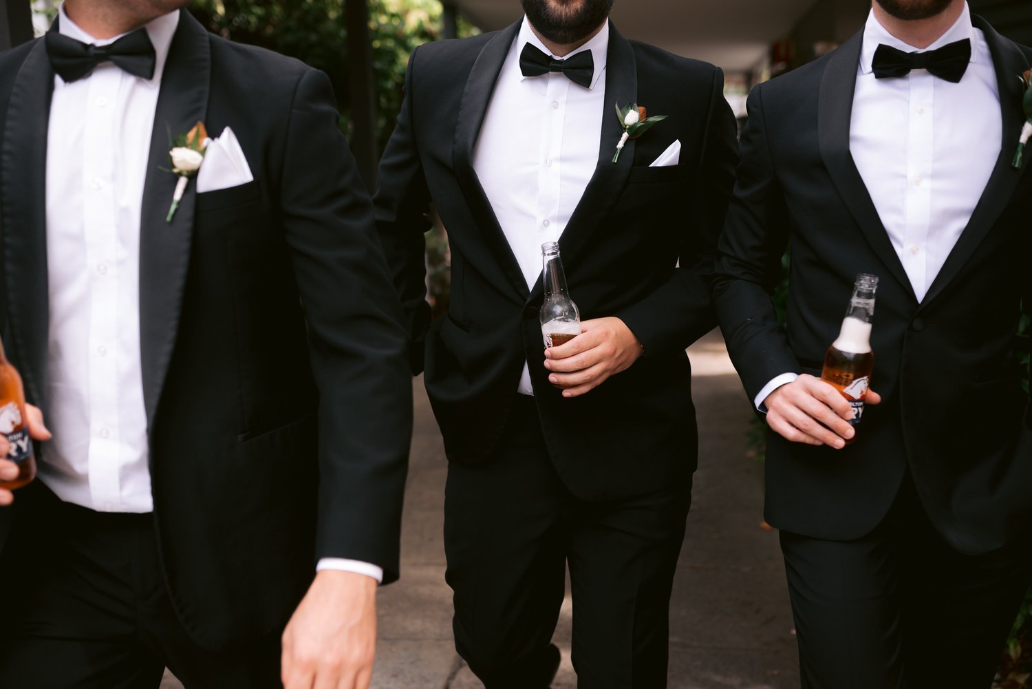 Three men in black suits and bow ties walking with bottles, likely groomsmen at a wedding.
