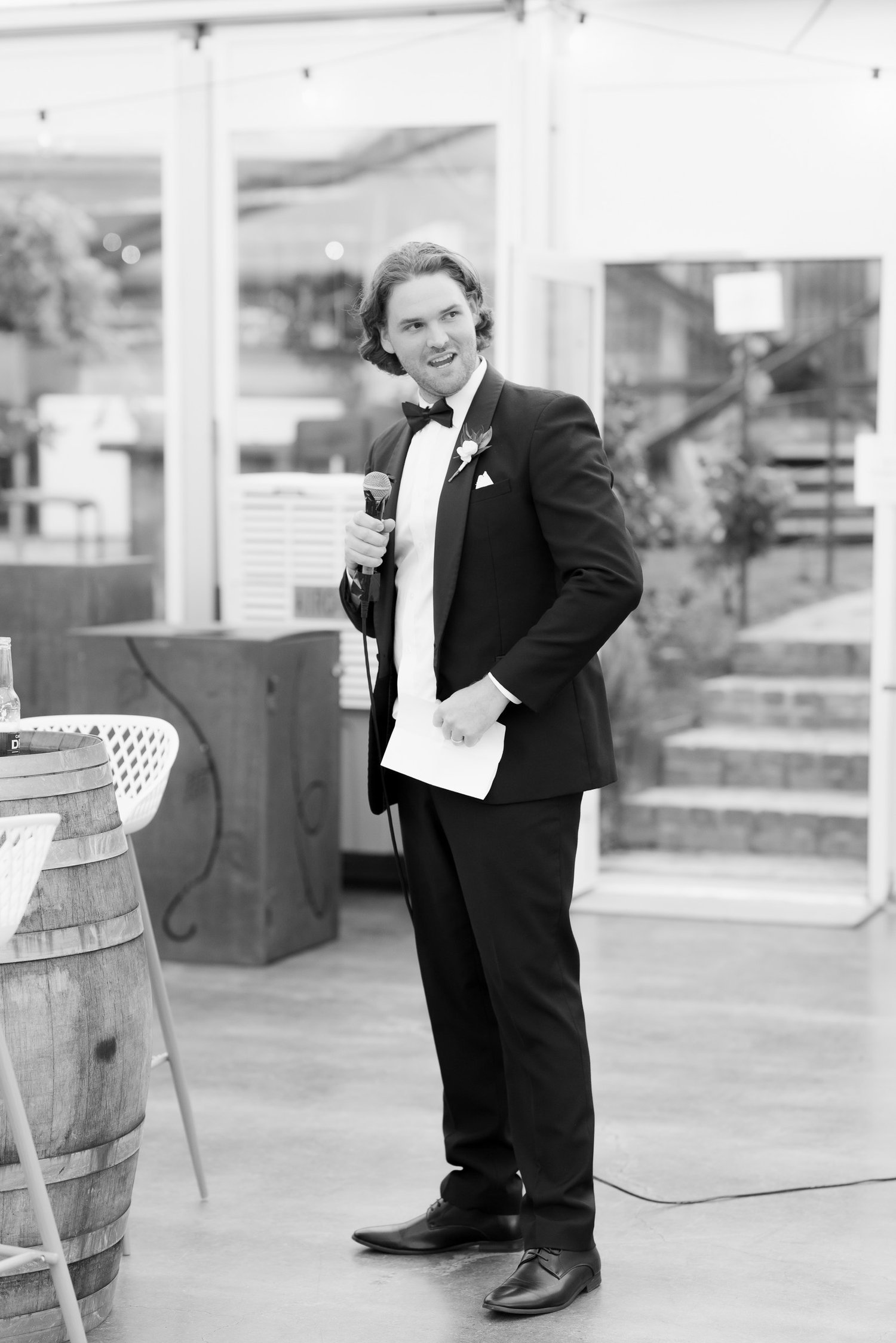 Man in a tuxedo giving a speech with a microphone, holding a paper, indoor event setting.