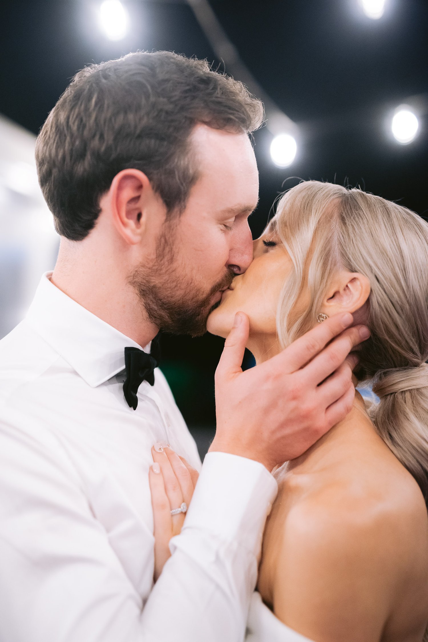A couple kissing at a wedding, with the groom wearing a bow tie and the bride showing her engagement ring. The background features soft, out-of-focus lights.