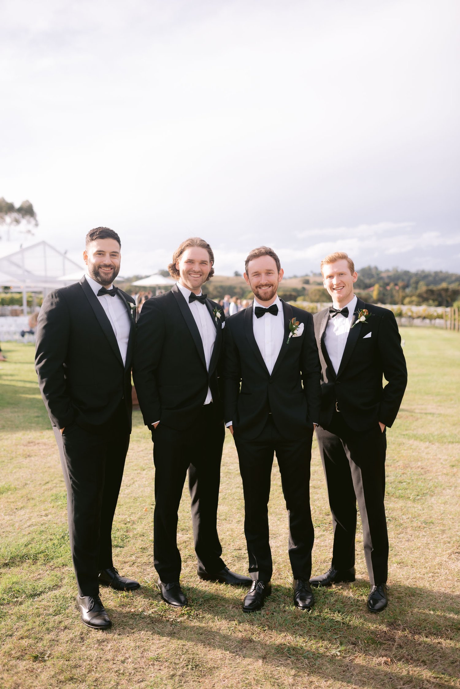 Four men in tuxedos standing on grass at an outdoor event.