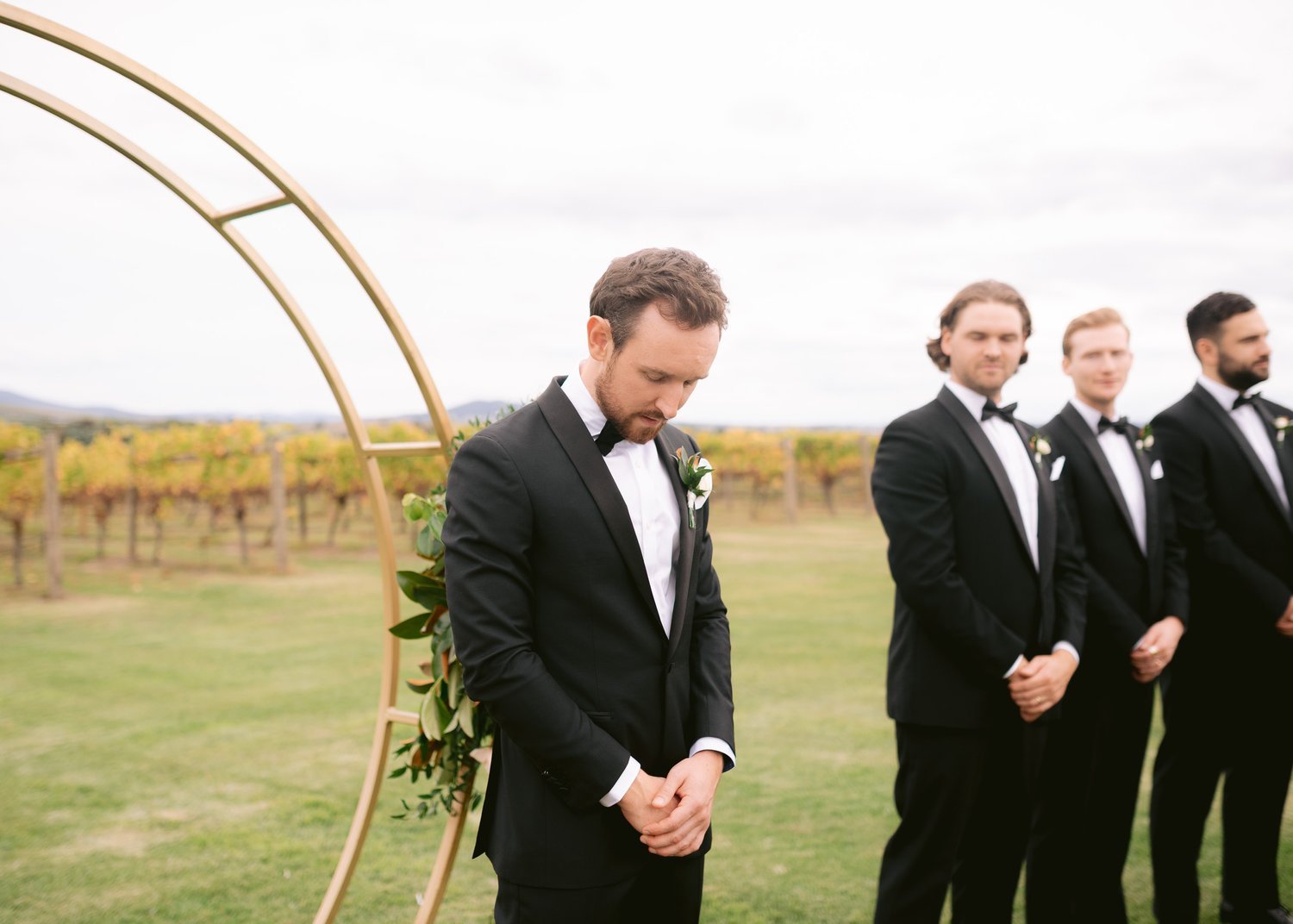 Outdoor wedding ceremony with groomsmen in black suits as groom waits excitedly for bride.