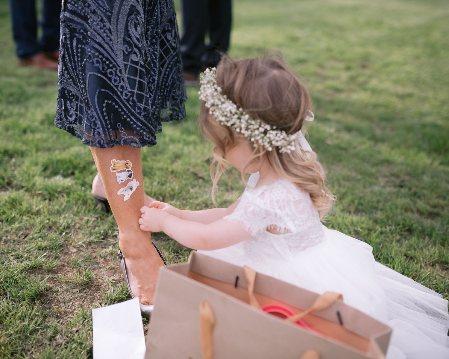 A young girl in a white dress applying stickers to a woman's leg during an outdoor event, with grass visible in the background.