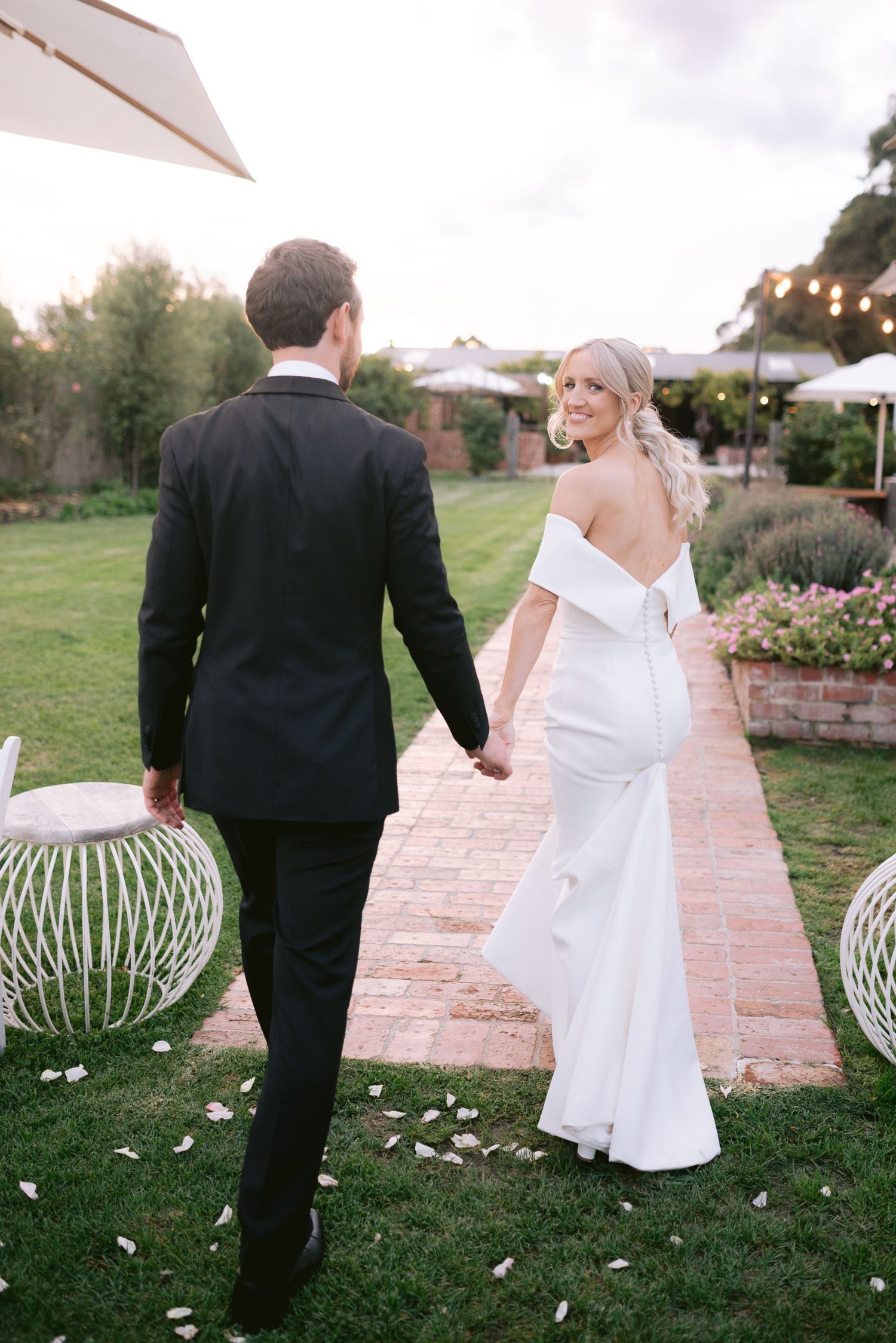 A newlywed couple walking hand in hand on a brick path in a garden. The bride is wearing a white wedding dress and the groom is in a black suit. The setting includes greenery, flowers, and outdoor seating.