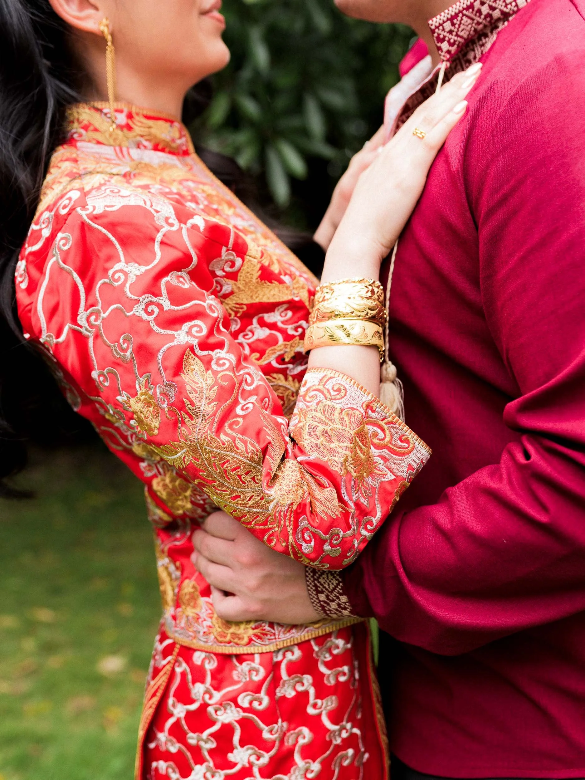 Close-up of a couple embracing, wearing traditional Asian outfits, with intricate red and gold embroidery, standing on grass.