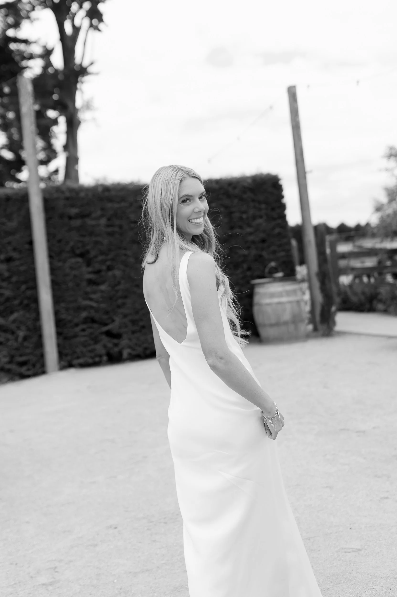 A woman in a white sleeveless dress smiling and looking over her shoulder outdoors with a hedge, trees, and a barrel in the background