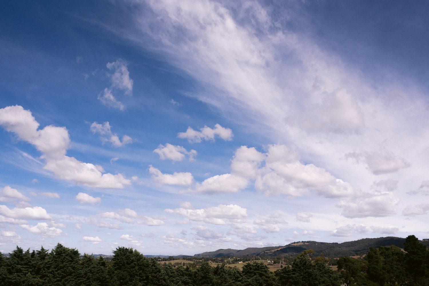 Panoramic landscape of Yarra Valley with a blue sky, white clouds, distant hills, and tree line.