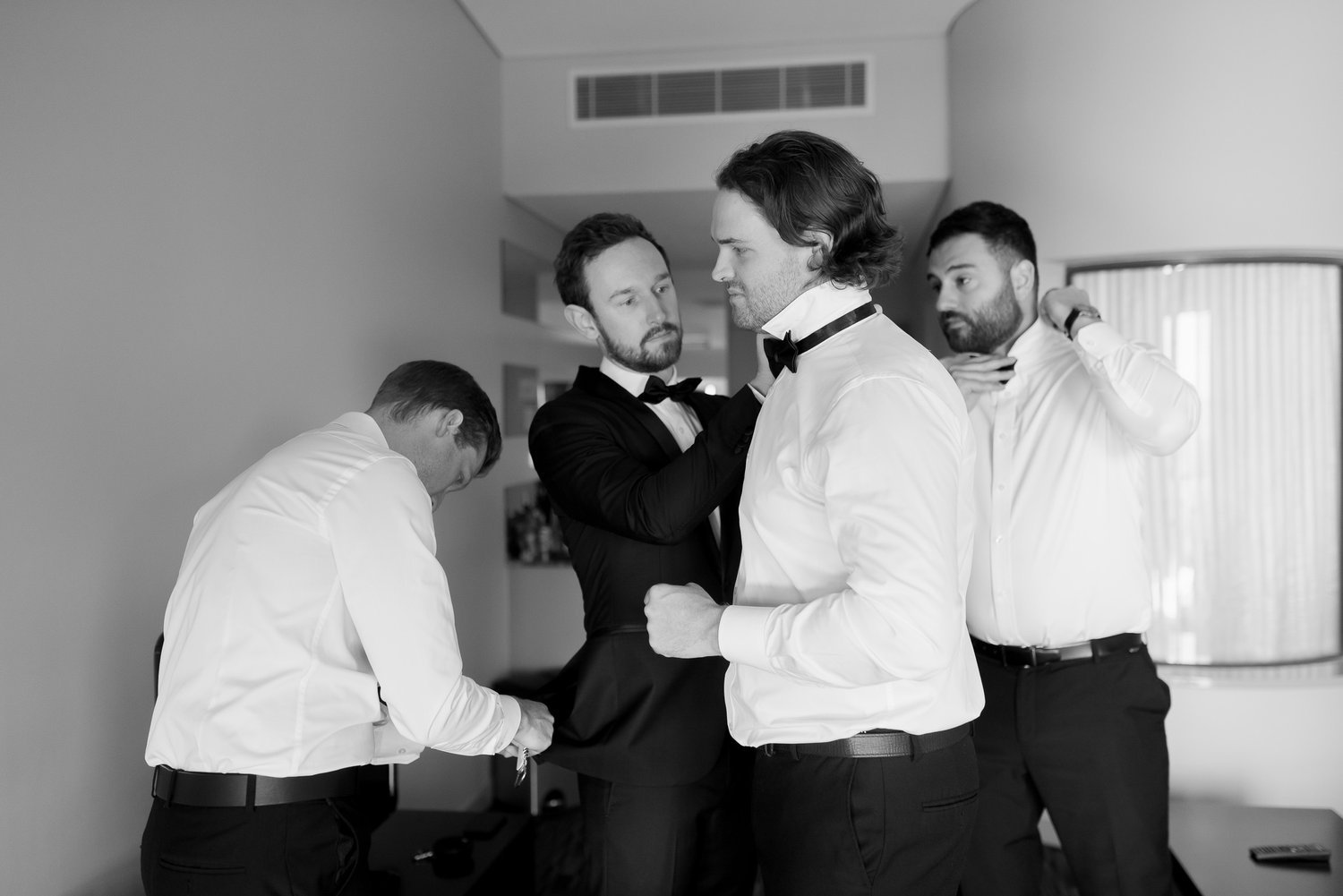 A group of four men in a room, all wearing formal attire. Three are adjusting each other's bow ties and suits, while the fourth is fixing his own tie. The image is in black and white.