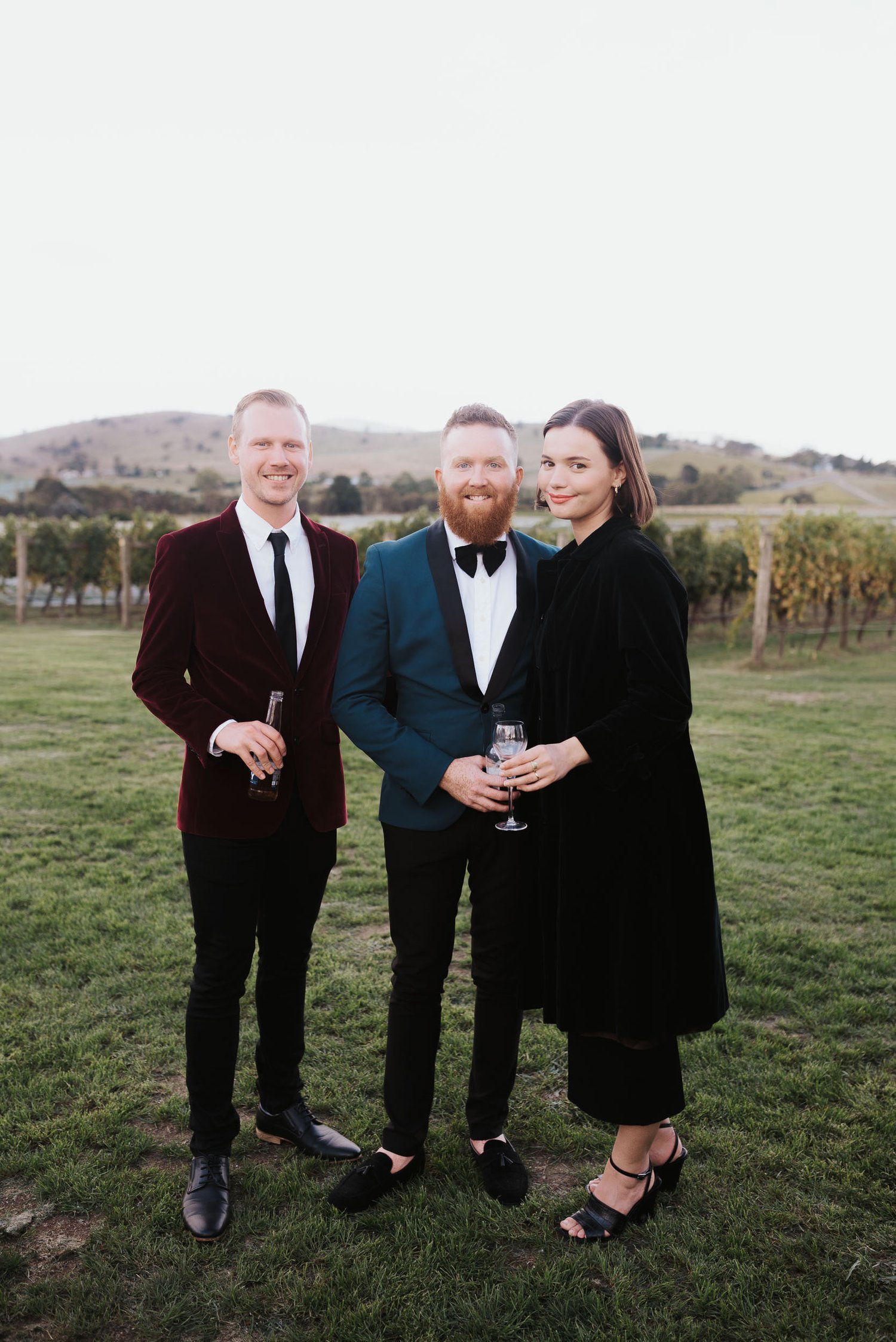 Three people dressed formally standing on grass, with rolling hills in the background.