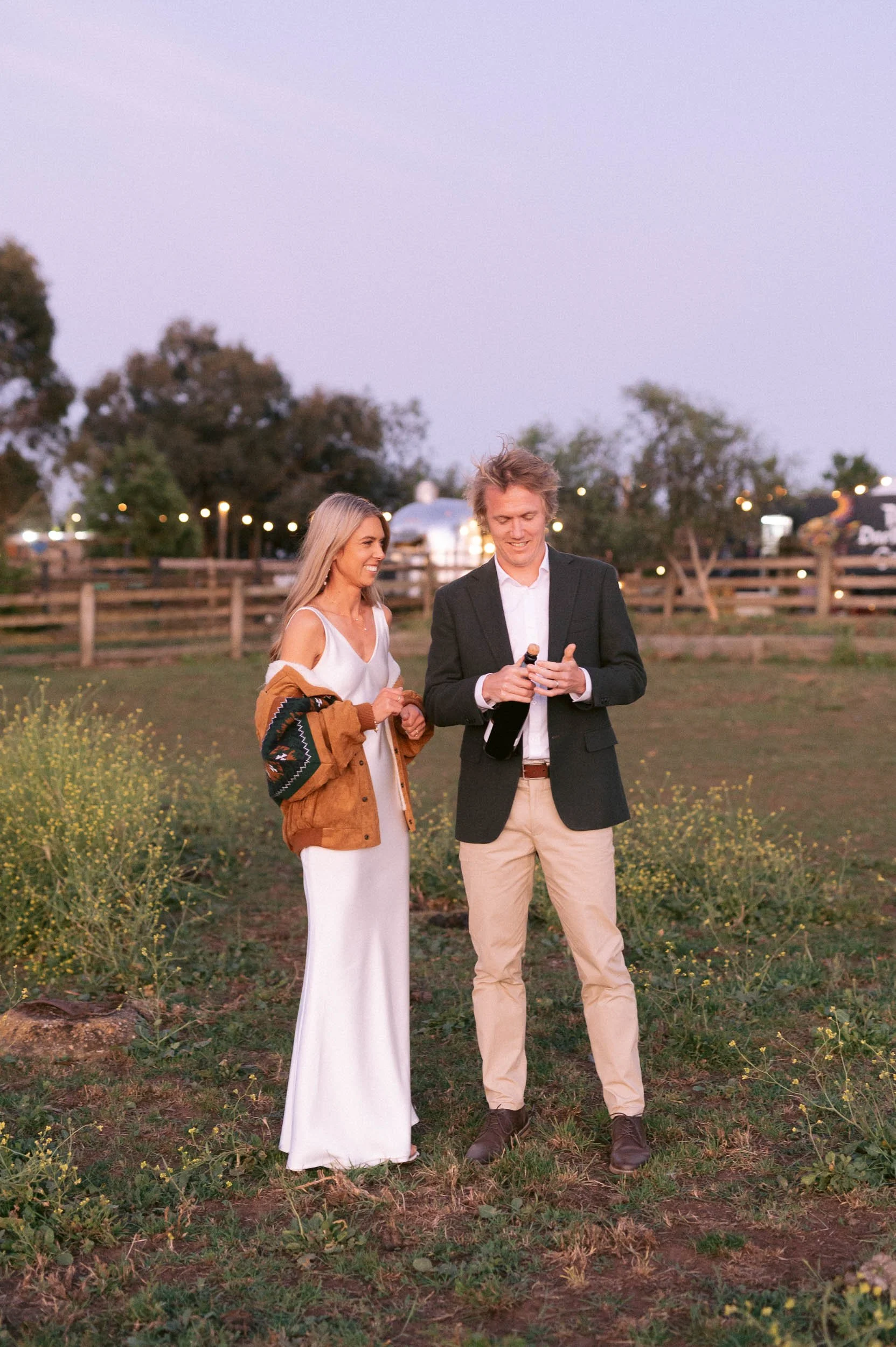 Newly weds standing outdoors on a grassy area with wildflowers, the woman in a white dress and brown jacket, the man in a black blazer and beige pants, holding a bottle of champagne, during sunset with a fence and trees in the background.