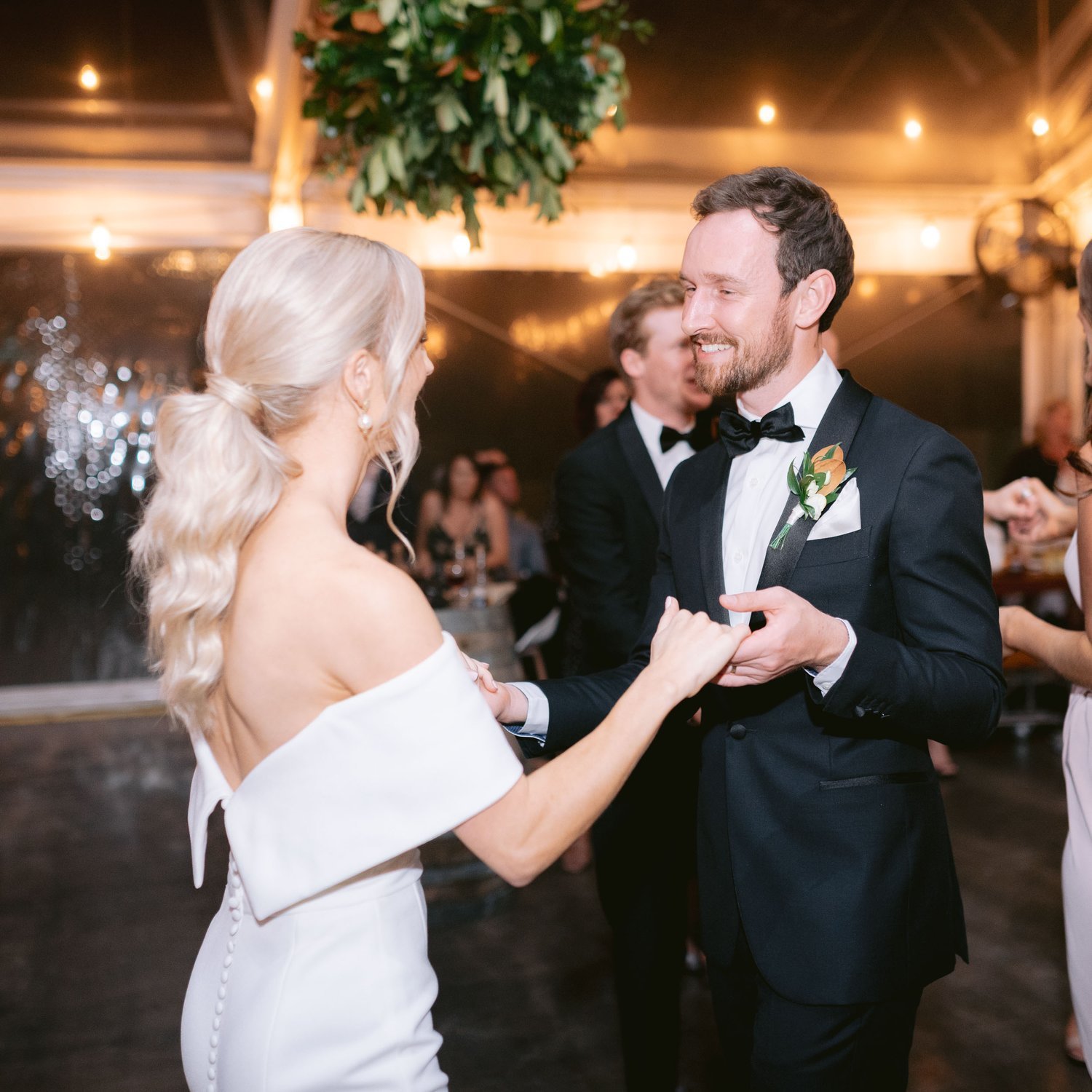 A bride and groom dancing at their wedding reception. The bride is wearing a white dress with an off-the-shoulder design and the groom is in a black tuxedo with a bow tie. They are inside a decorated venue with hanging greenery and lights.