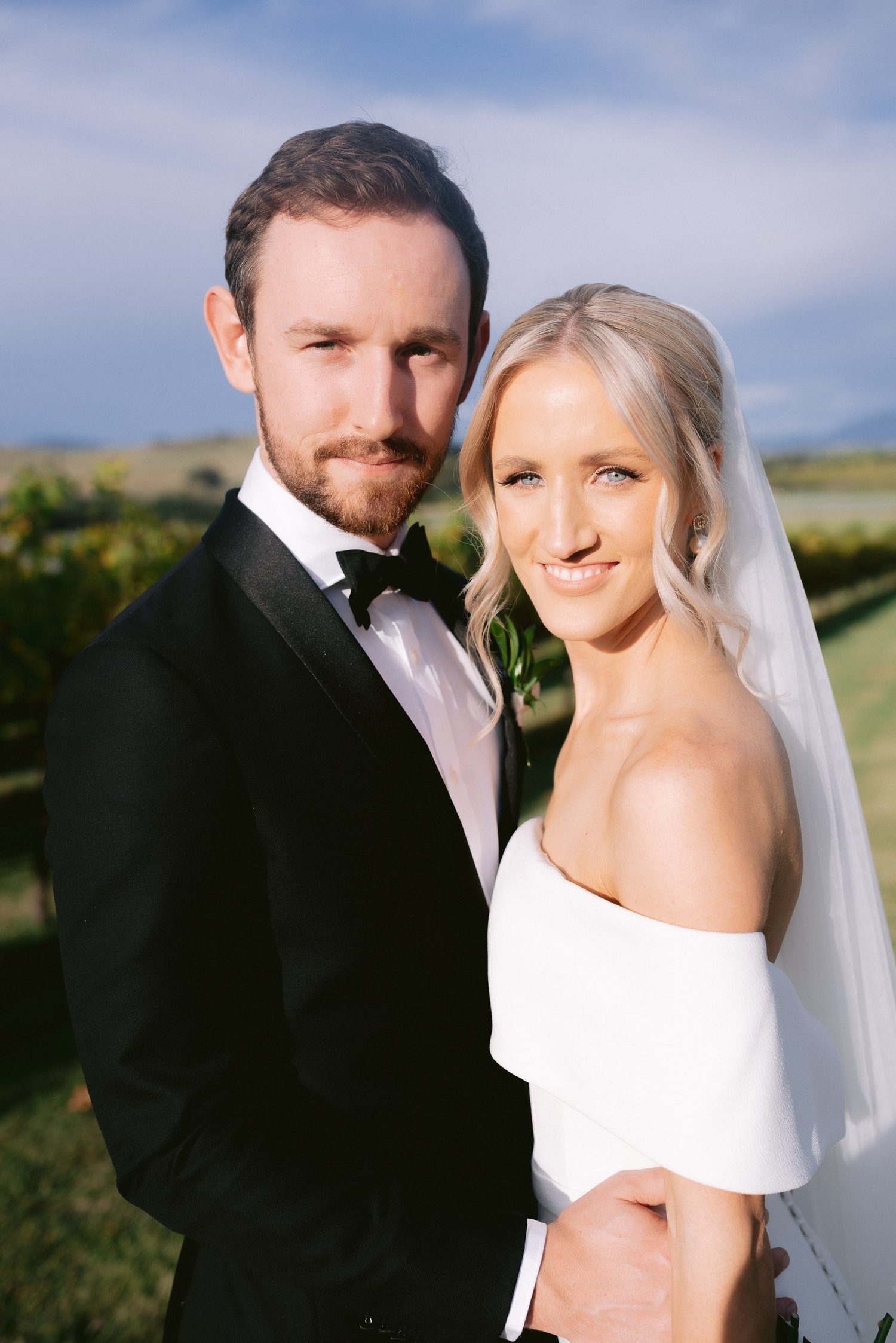 A bride and groom in wedding attire posing outdoors. The groom is in a black tuxedo, and the bride is in a white off-the-shoulder dress with a veil.