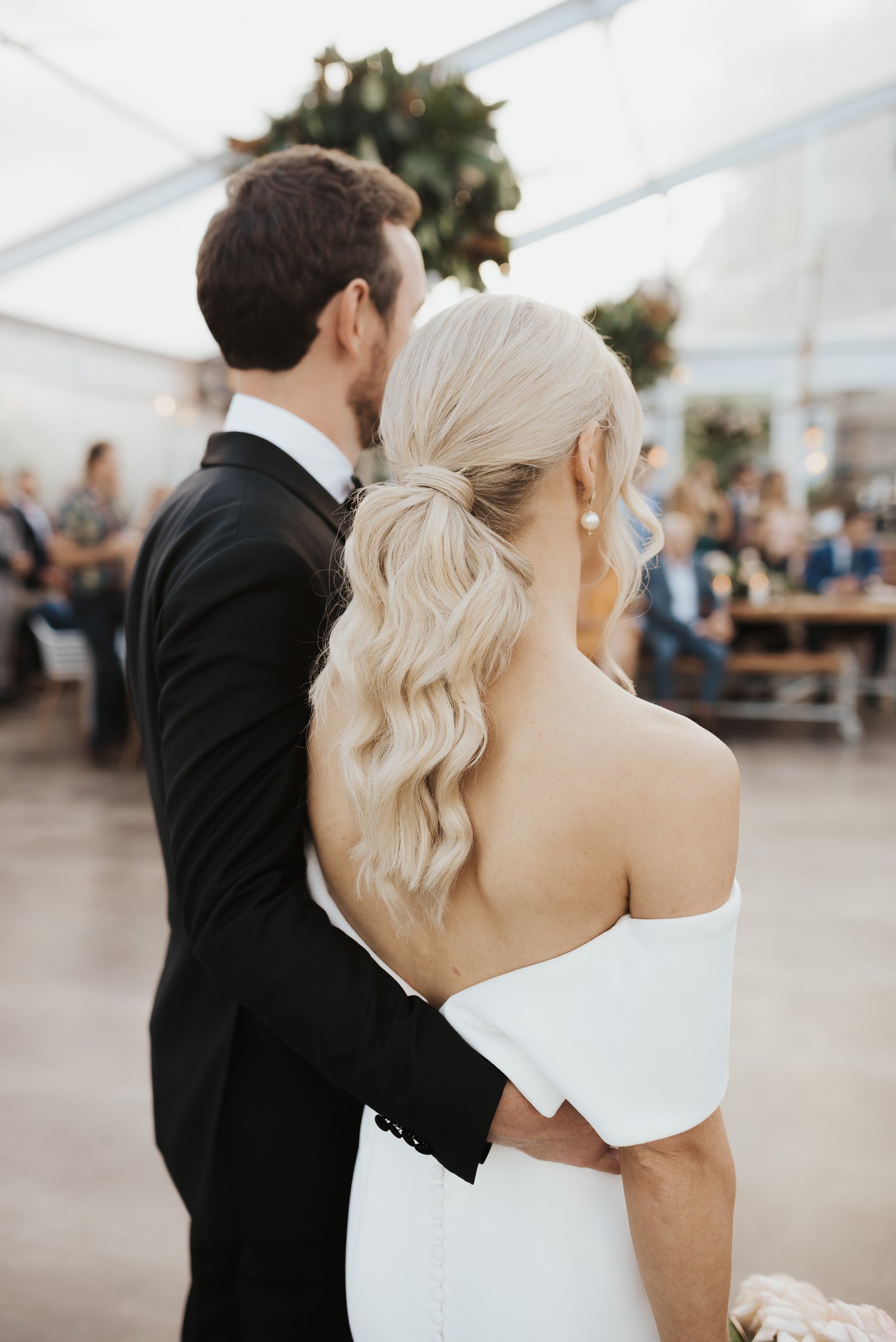 Bride and groom embracing at a wedding, with the bride in a white gown and the groom in a black suit, surrounded by blurred guests.