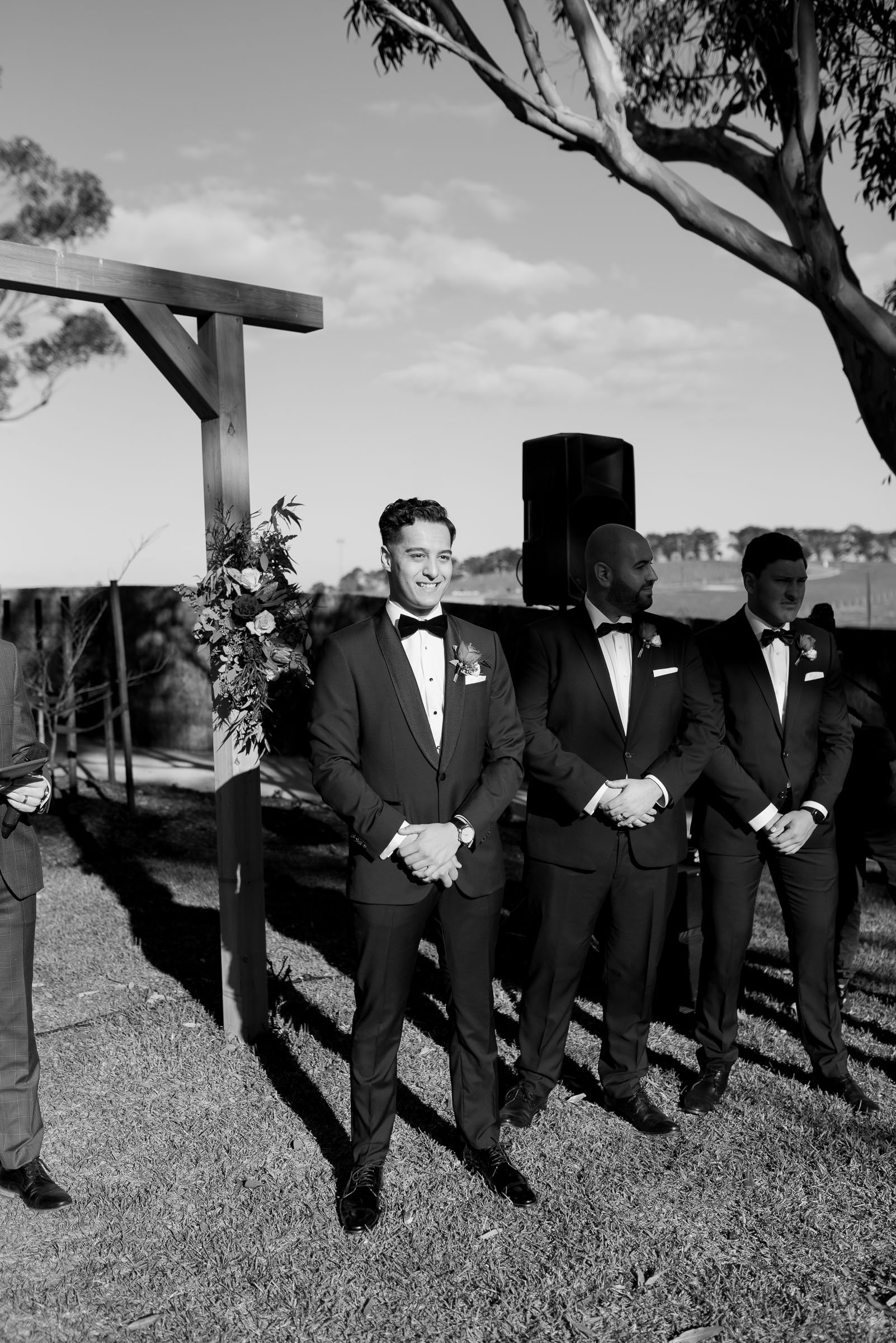 Groomsmen in suits standing outdoors at a wedding ceremony under a wooden arch with floral decoration.