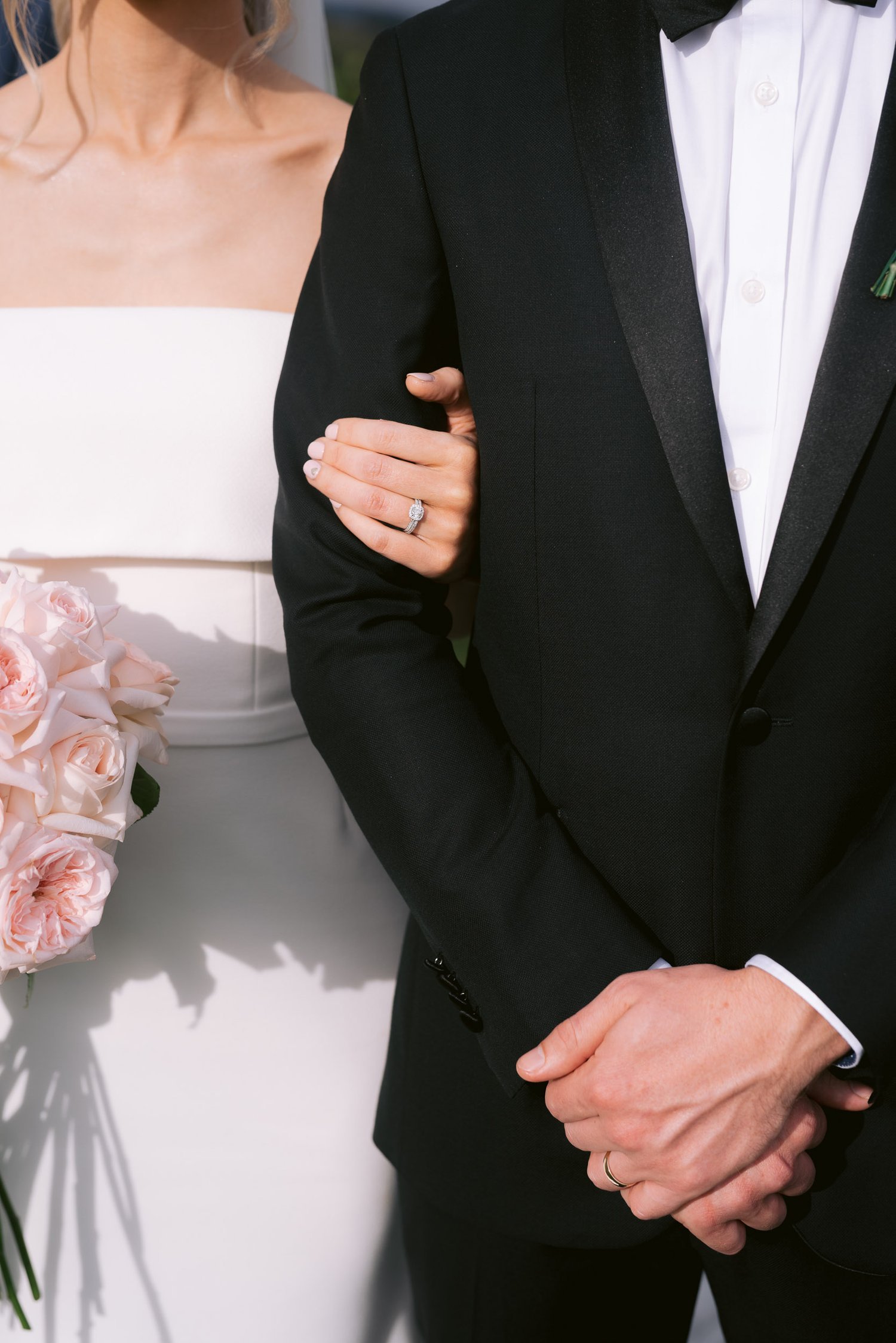 Bride and groom standing together, bride holding pink rose bouquet, wearing wedding dress, groom in black tuxedo.