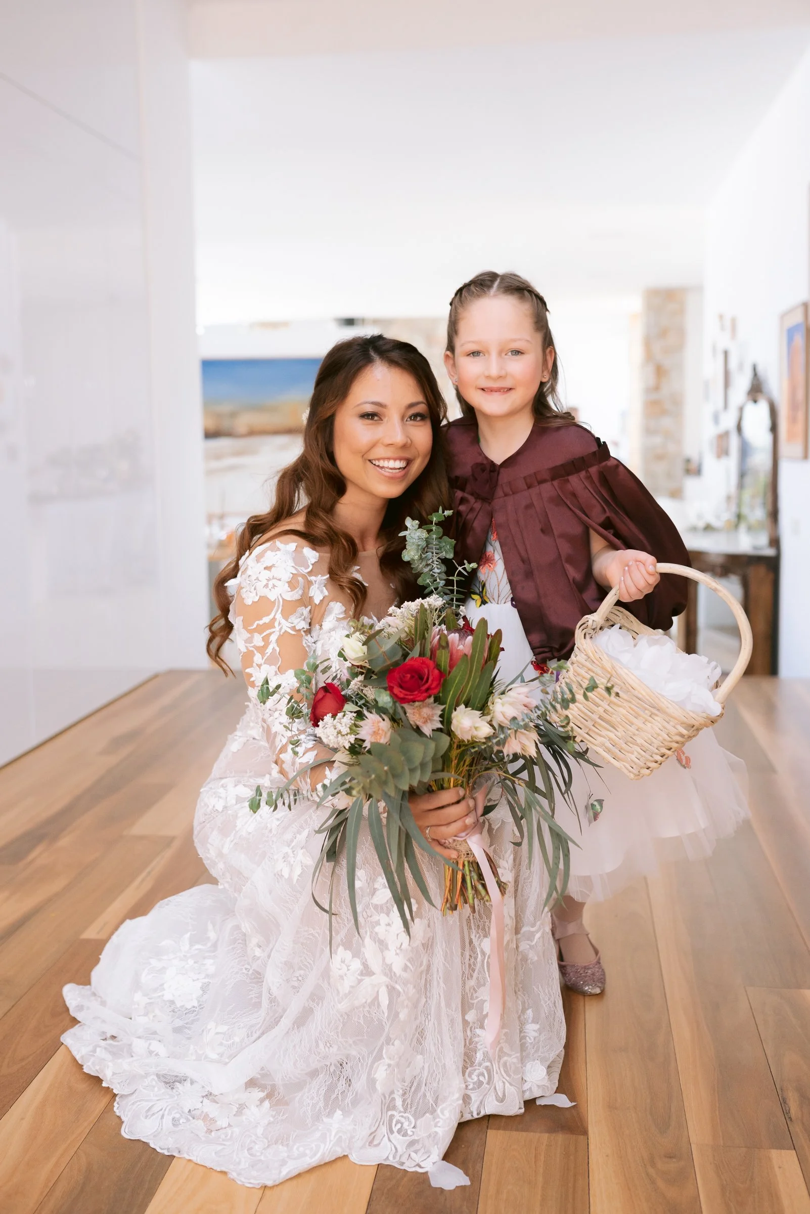 Bride in lace dress holding bouquet, posing with a young girl in a maroon cape holding a wicker basket.