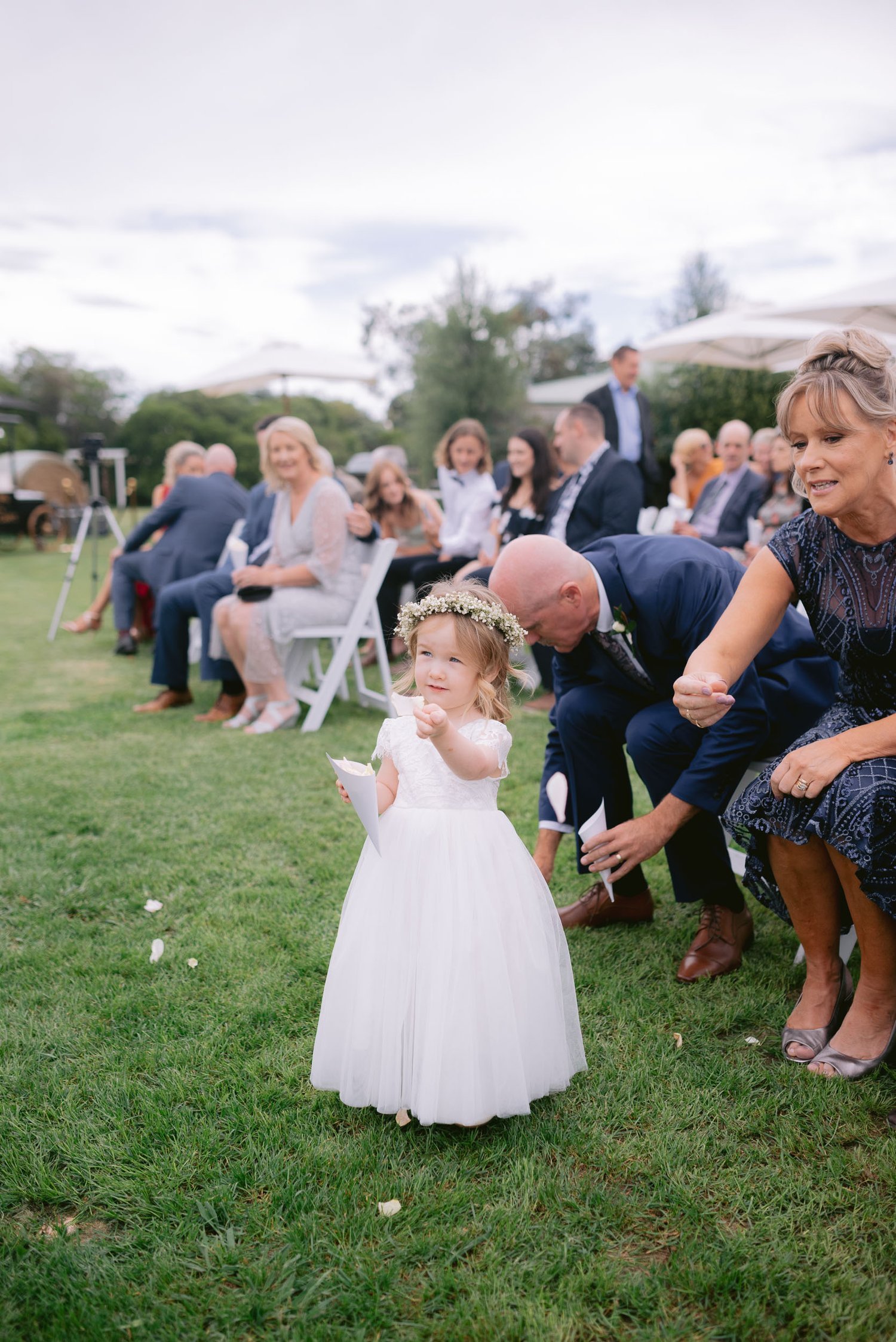 Young flower girl in a white dress with a floral headband, holding petals, standing on grass, surrounded by seated wedding guests in an outdoor setting.