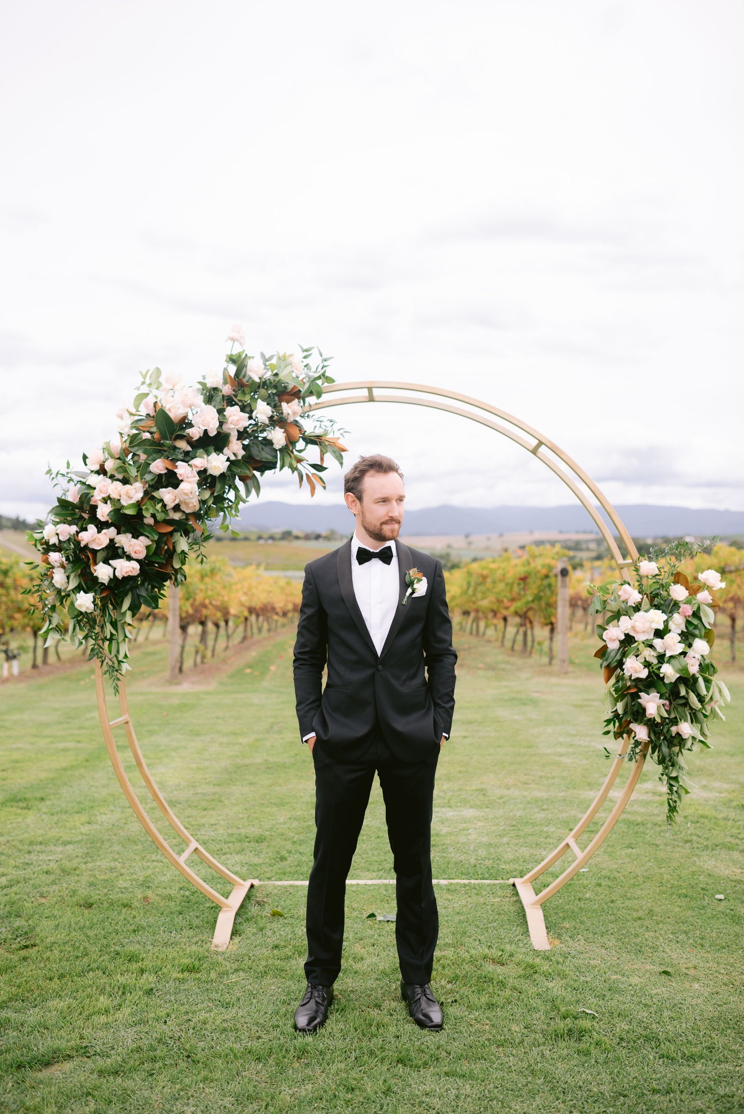 Man in black suit and bow tie standing in front of a circular floral arch on a grassy vineyard setting.