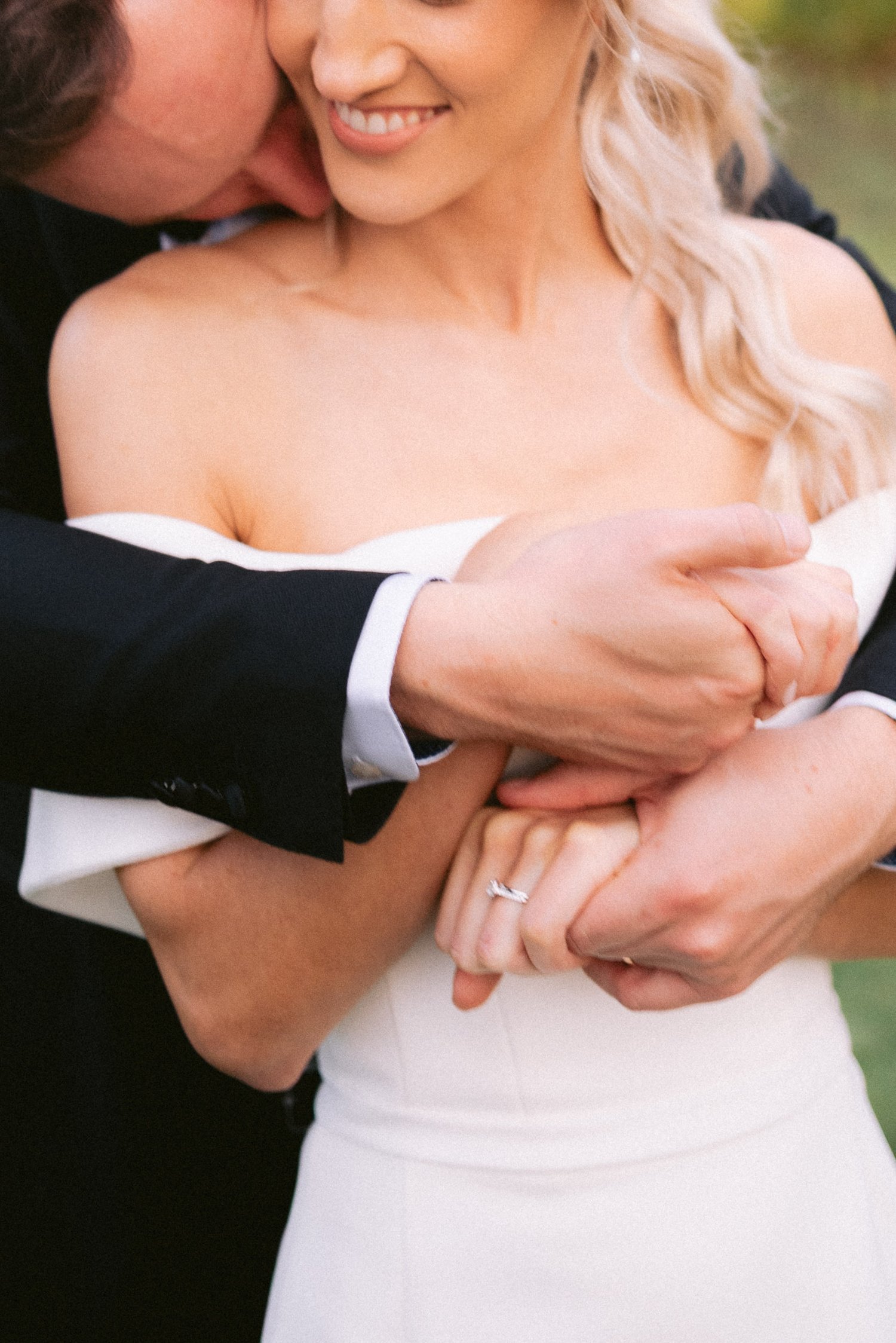 Close-up of a couple embracing, bride in a white dress showing engagement ring, groom in a black suit.