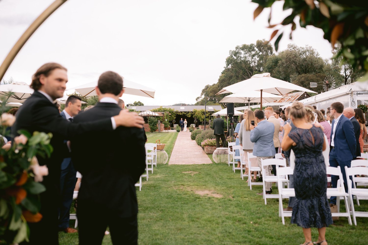 Outdoor wedding ceremony with guests seated in white chairs, a groom and groomsman in suits standing on the left, and a bride walking down an aisle lined with grass and bricks.