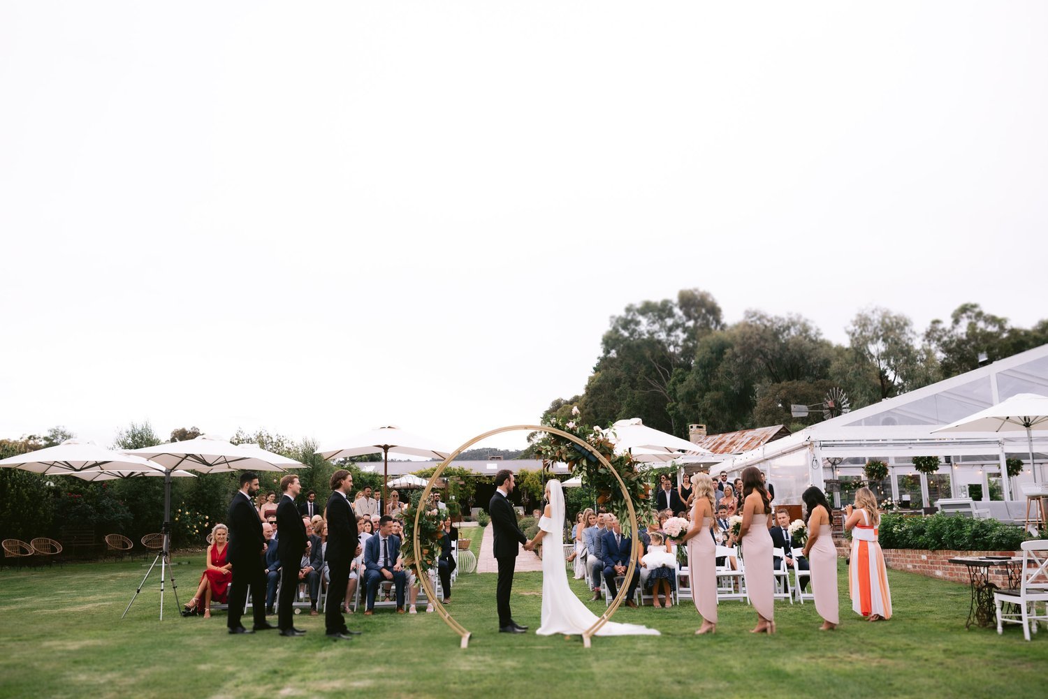 Outdoor wedding ceremony with a couple standing under a floral arch, surrounded by bridesmaids, groomsmen, and guests seated on the lawn. White umbrellas and a tent are in the background.