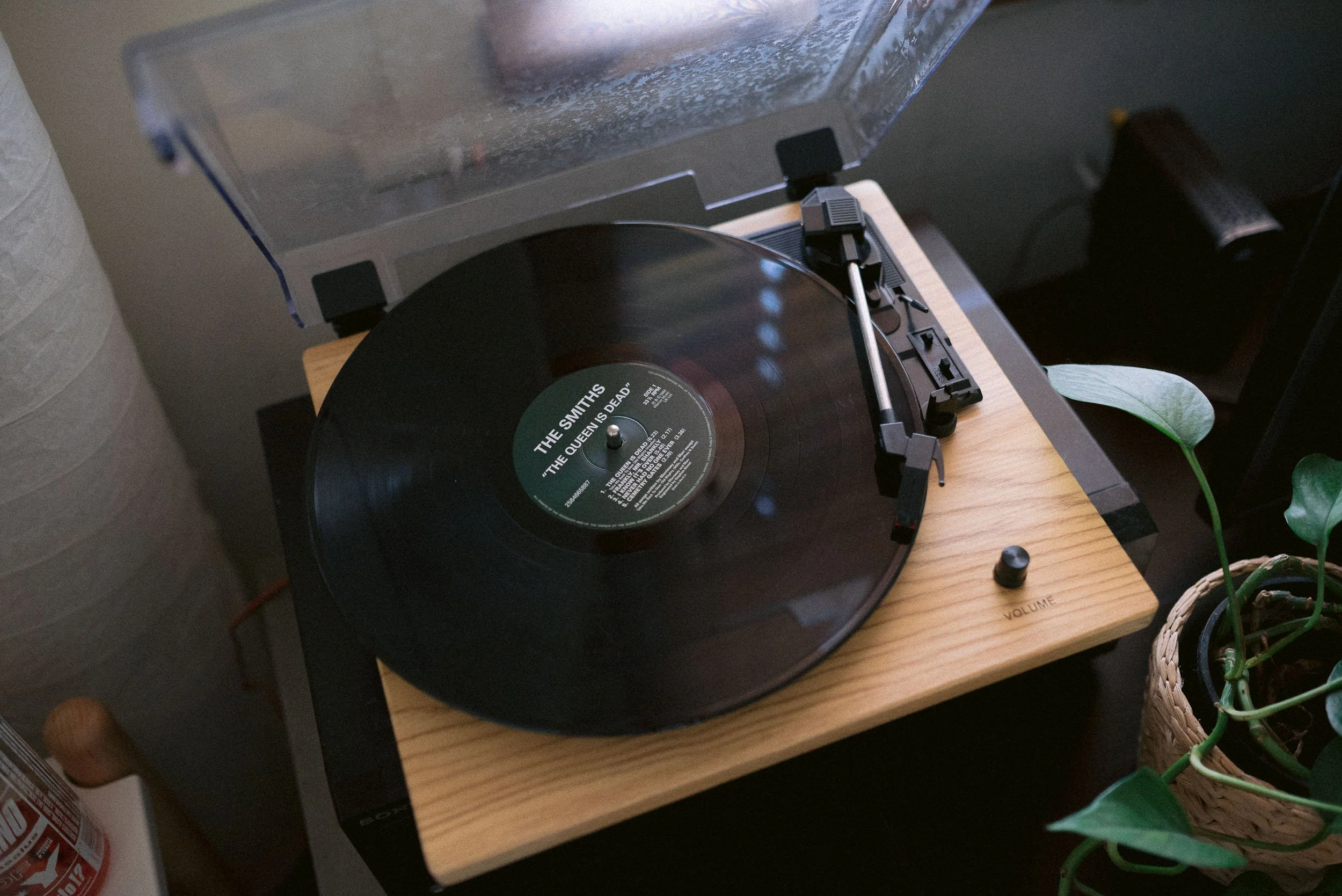 A turntable playing The Smiths' vinyl record 'The Queen Is Dead' on a wooden stand, with a potted plant nearby.