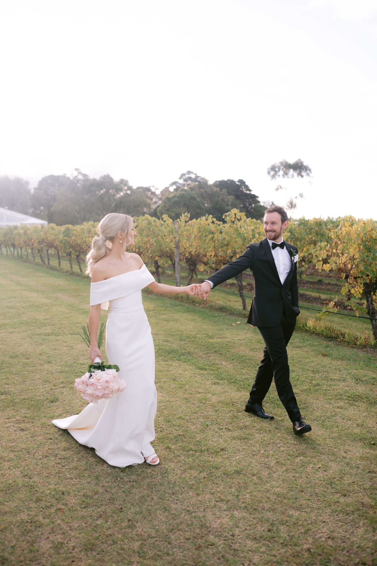 Bride and groom walking hand in hand through a vineyard, bride holding pink flowers, in a white wedding dress and groom in black suit with bow tie.