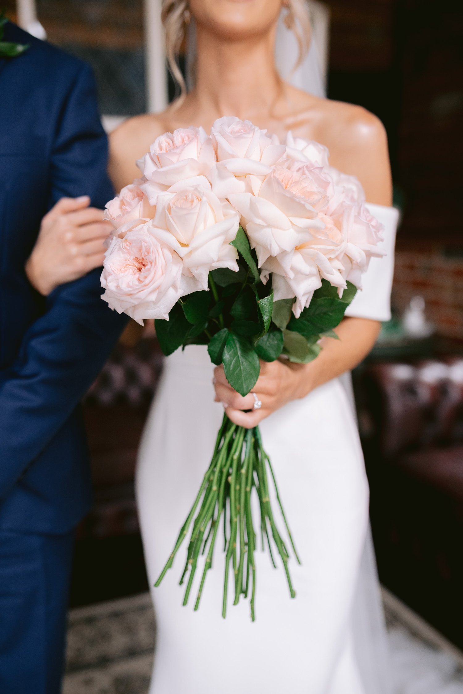 Bride holding a bouquet of light pink roses, wearing a white dress and an arm of a person in a blue suit is visible beside her.