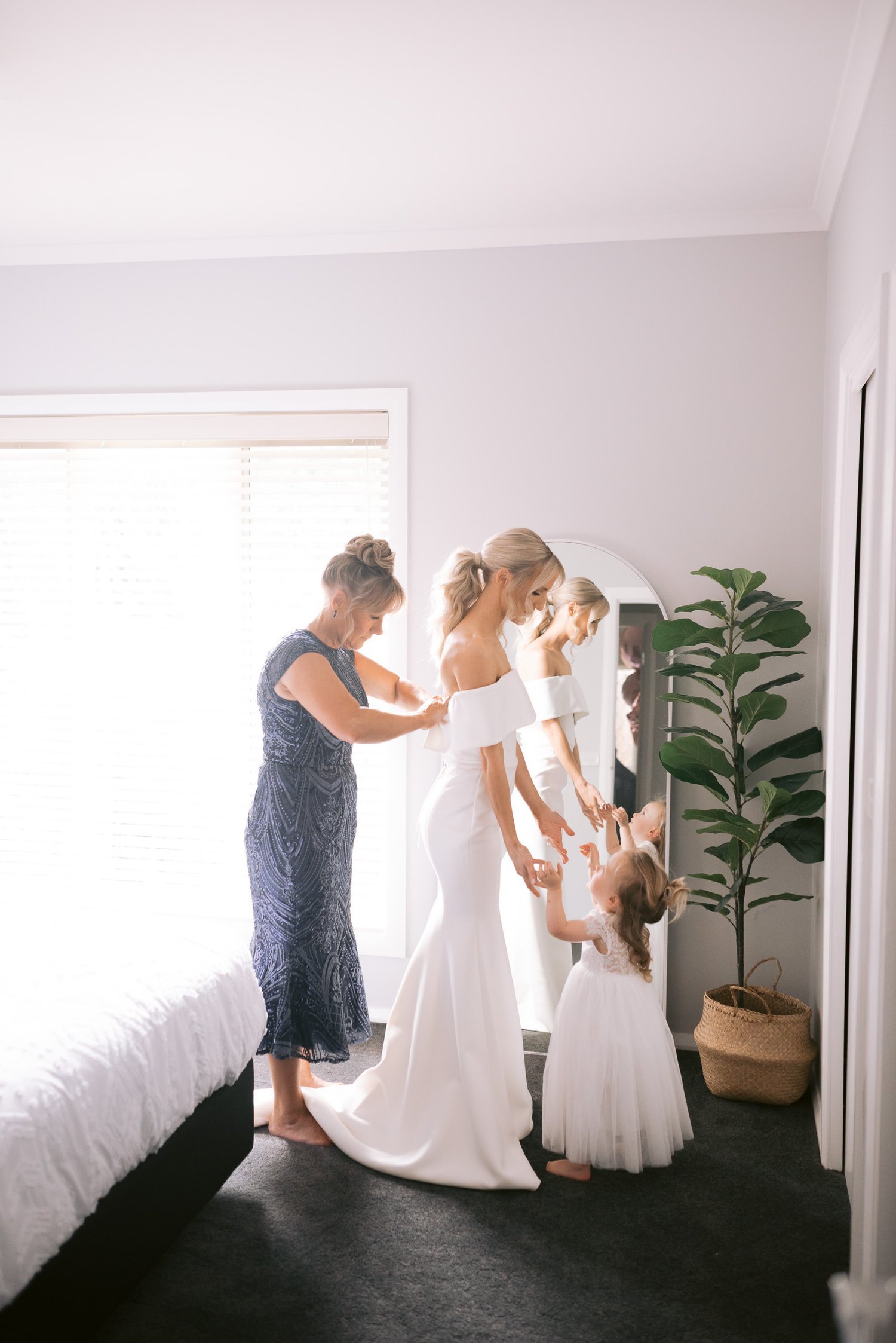 A bride in a white gown being assisted by an older woman, while a young girl in a white dress stands nearby in a bedroom with a mirror and potted plant.
