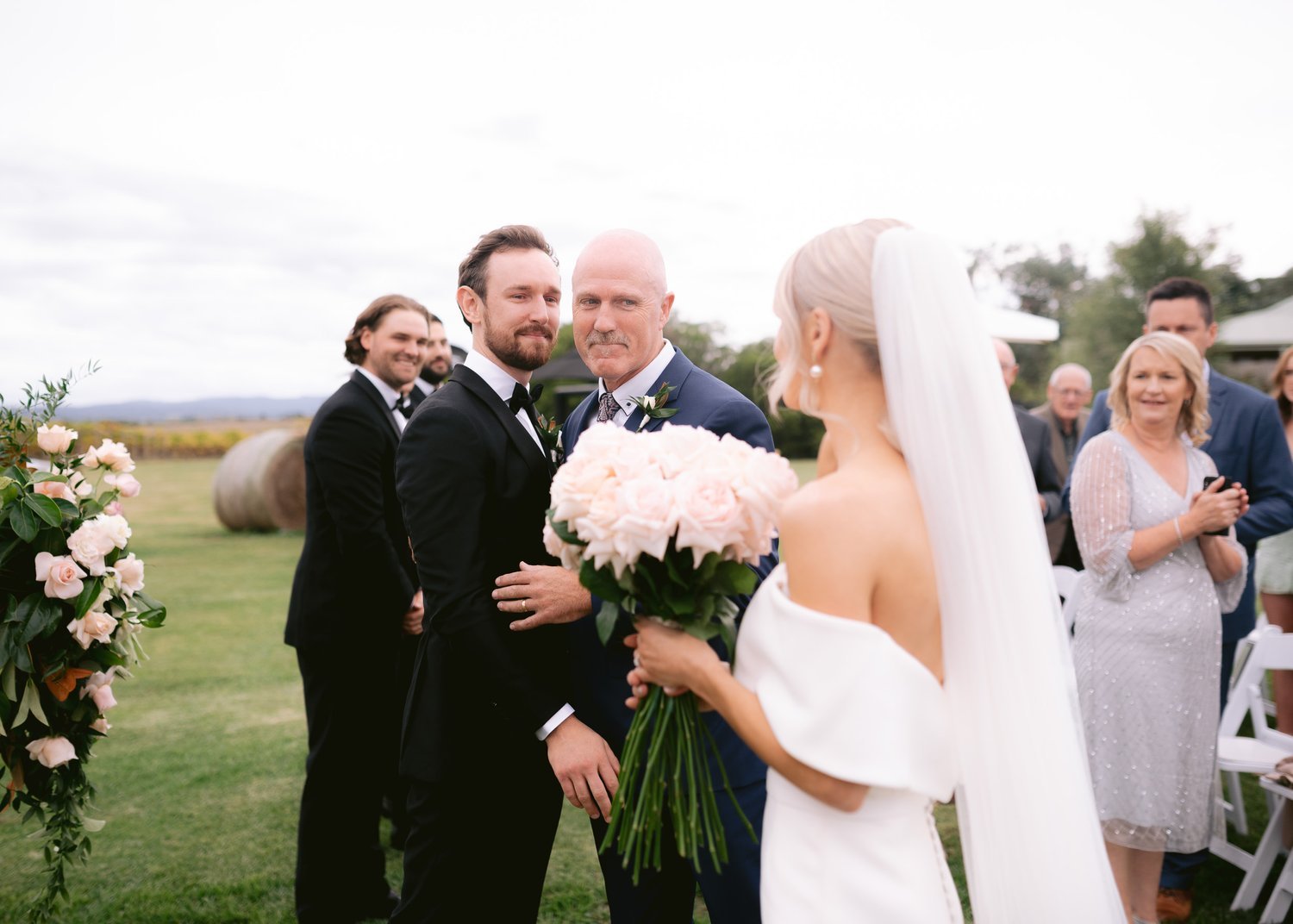 Outdoor wedding ceremony with bride holding pink bouquet, groom in black suit embracing older man, guests in formal attire seated in background.