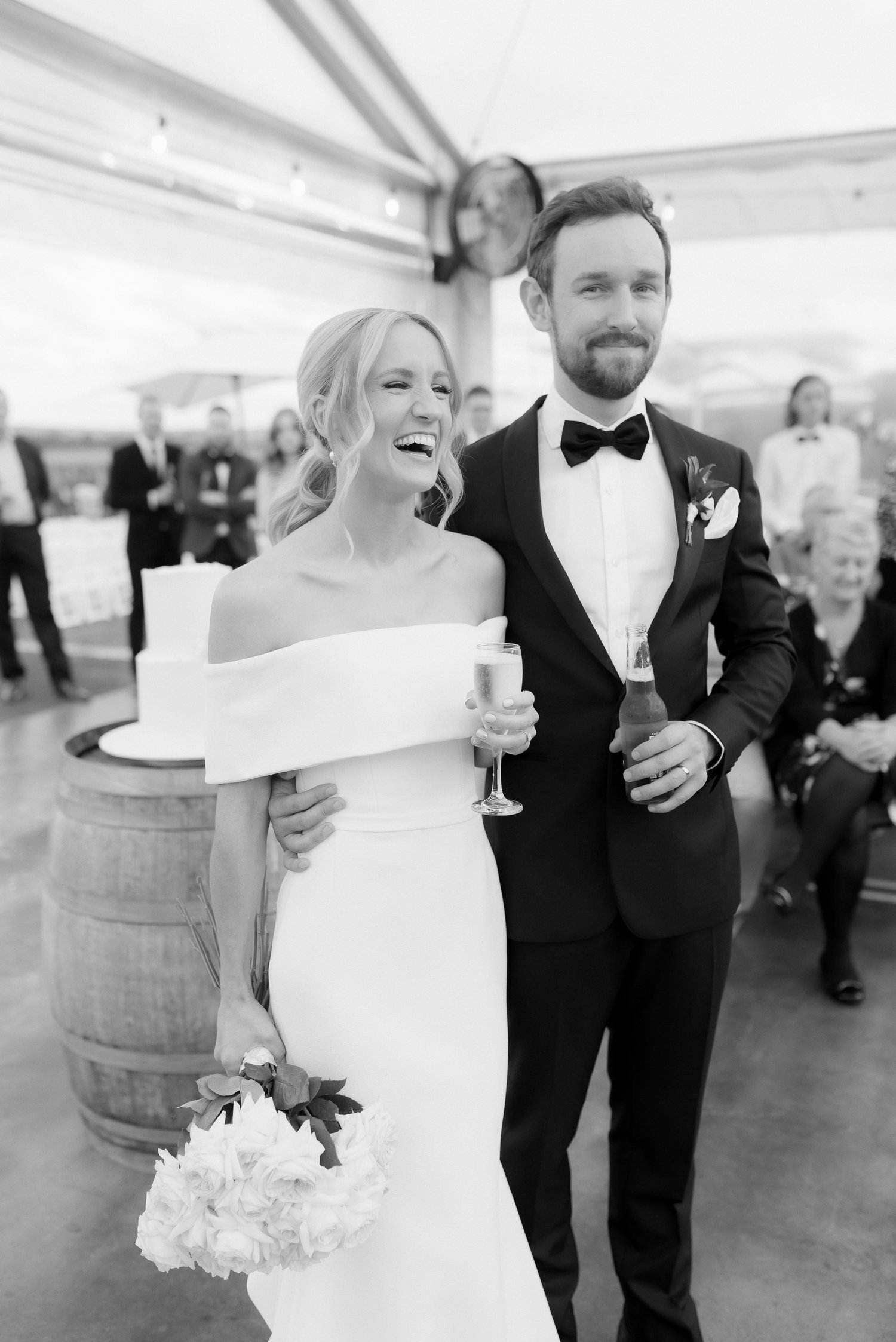 Smiling bride and groom celebrating at wedding reception, with the bride holding a bouquet and a champagne flute, and the groom holding a beer. They are in a tented venue, surrounded by guests.