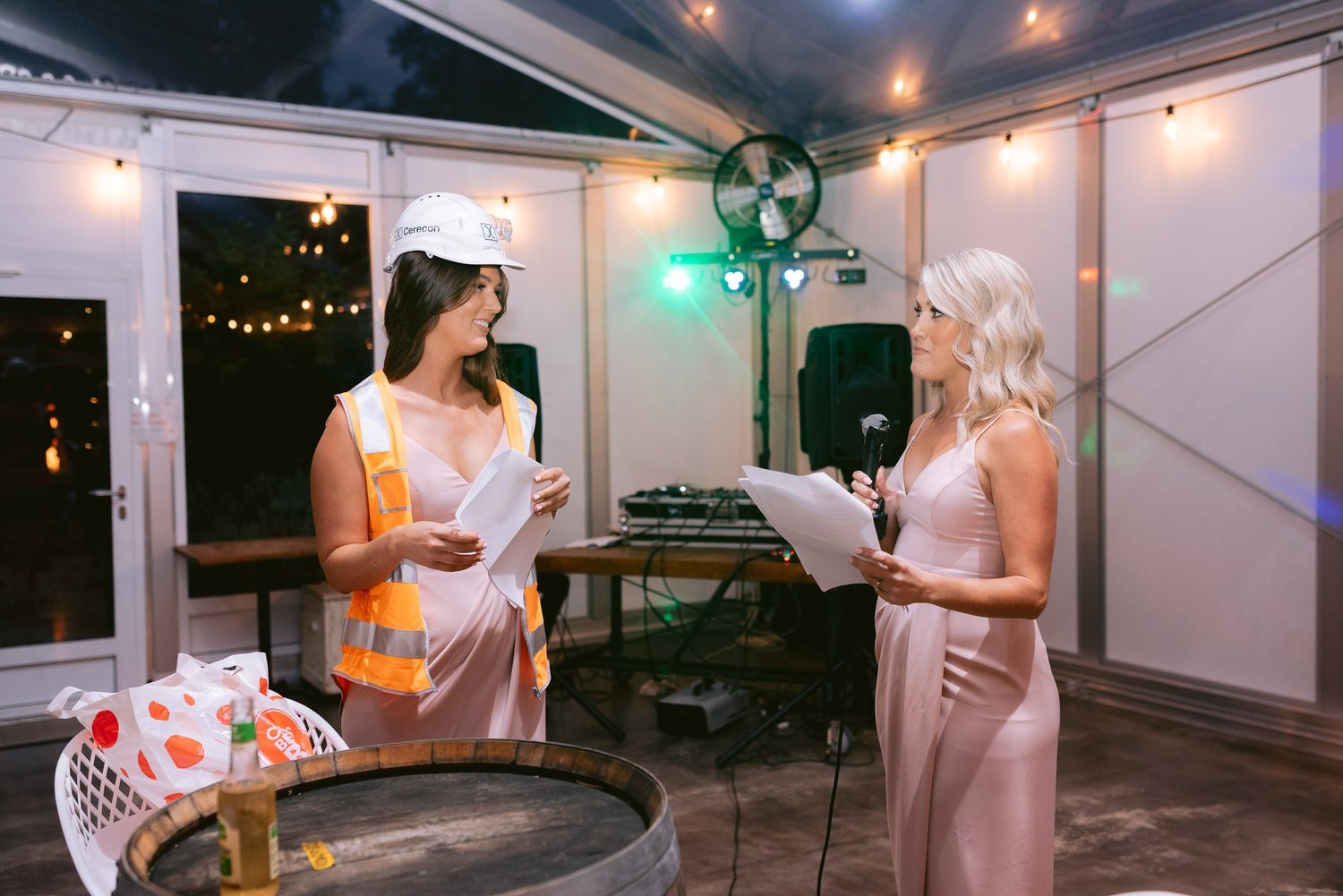 Two women dressed in formal attire, one wearing a hard hat and reflective vest, holding papers and speaking at an indoor event with string lights and DJ equipment in the background.