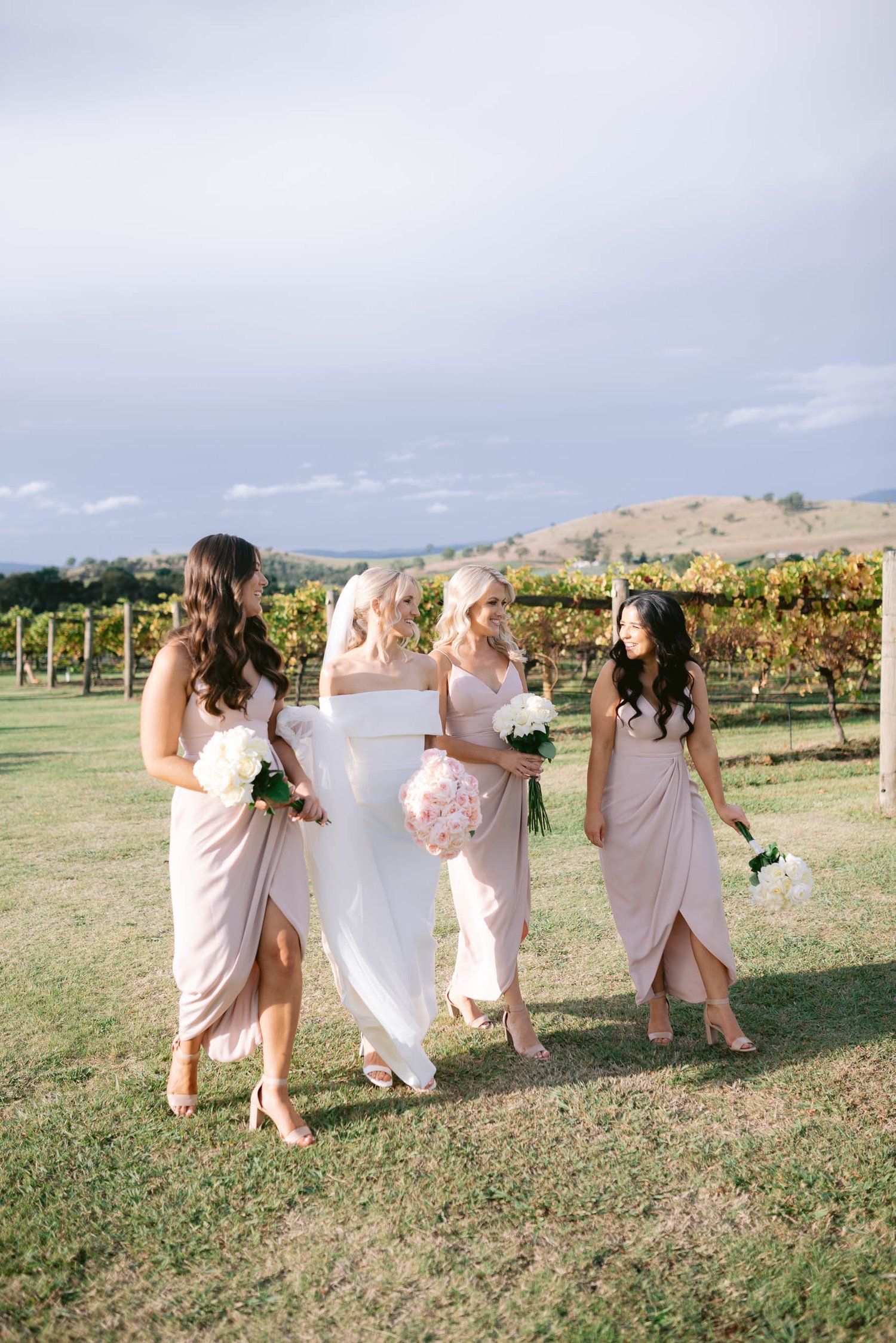 A bride in a white gown with three bridesmaids in blush pink dresses walking outdoors, holding bouquets, with a vineyard and hills in the background.