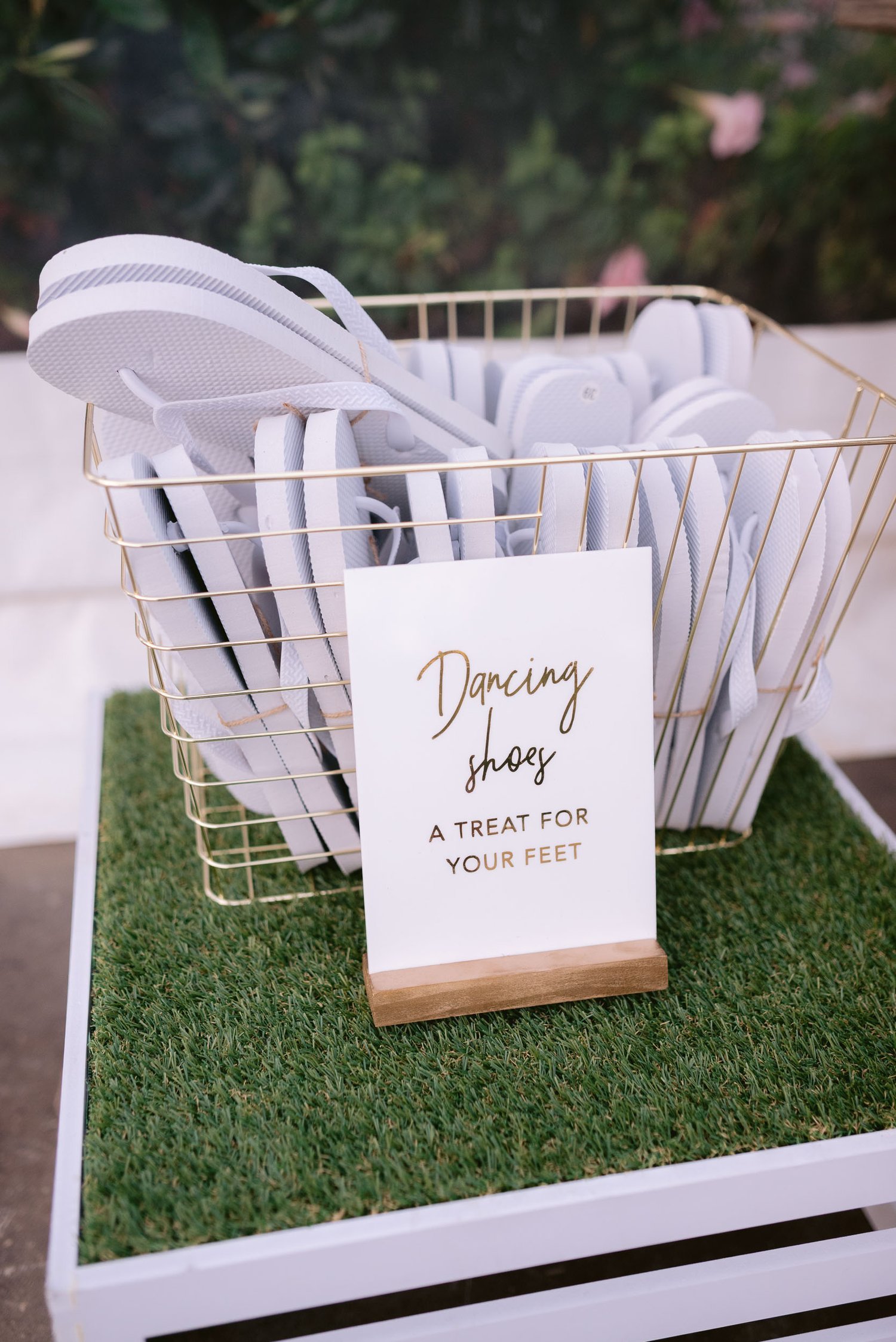 Wire basket filled with white flip-flops labeled "Dancing shoes, a treat for your feet" on a grass-like mat.