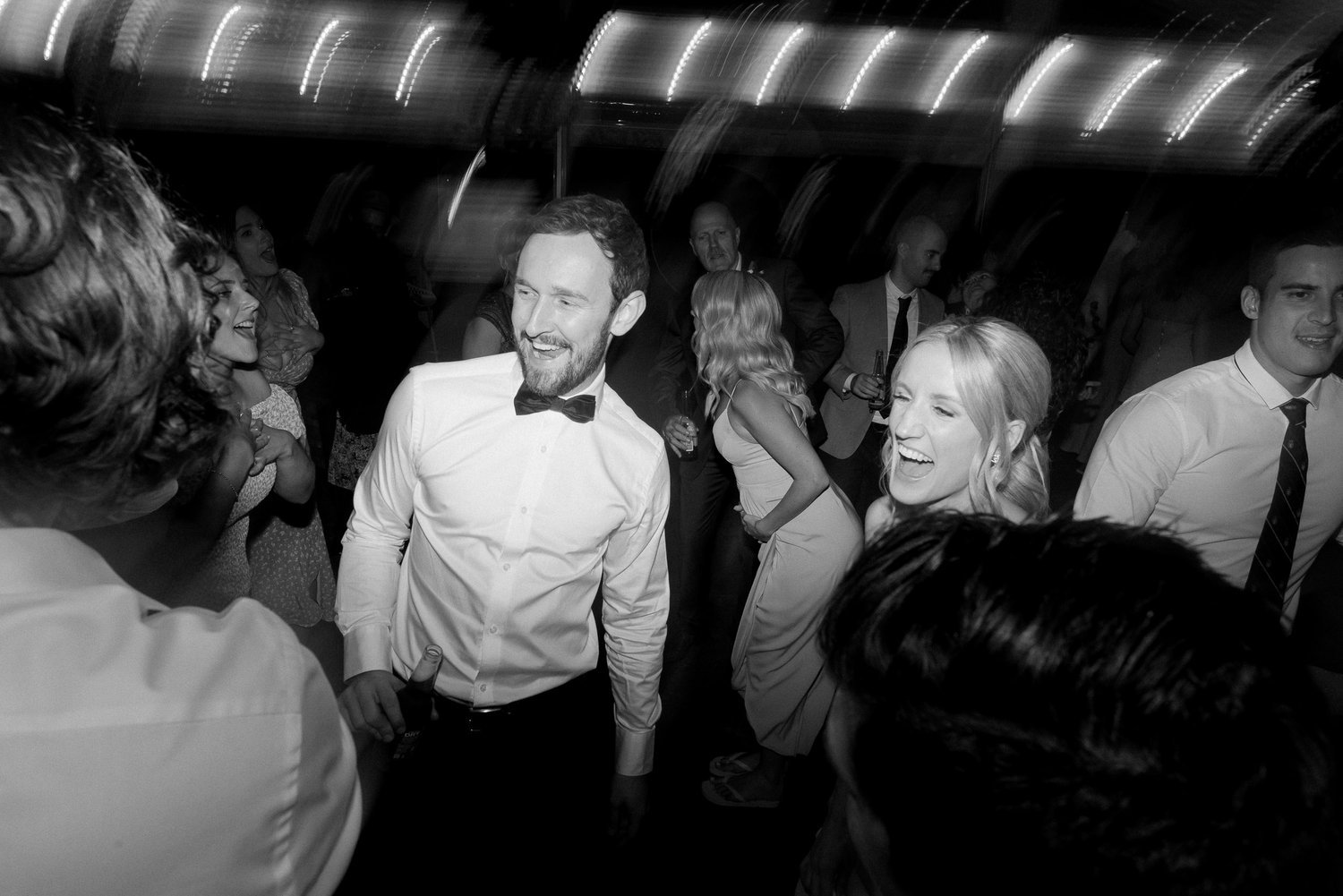 Black and white photo of a group of people dancing at a party with lights above.