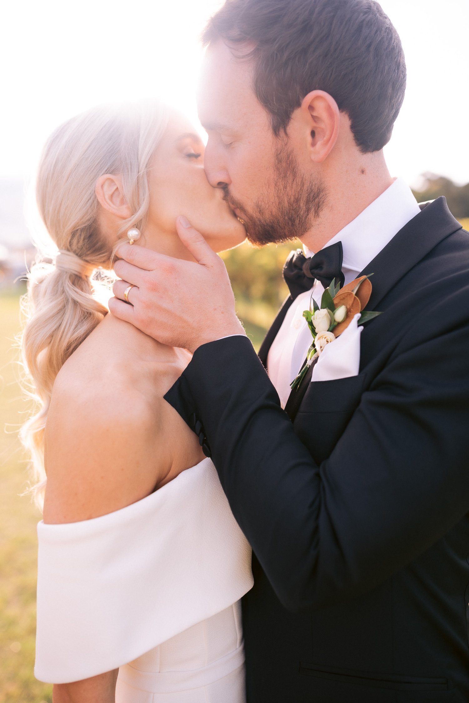 Bride and groom kissing outdoors in sunlight, wearing wedding attire.