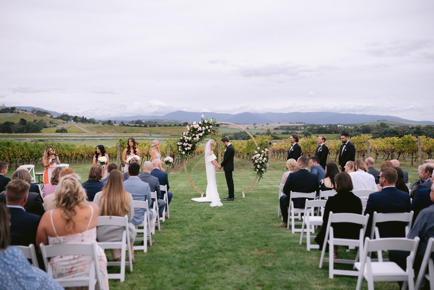 Outdoor wedding ceremony in a vineyard with bride and groom under a floral arch, surrounded by guests seated in white chairs, bridesmaids, and groomsmen. Scenic landscape with mountains in the background.