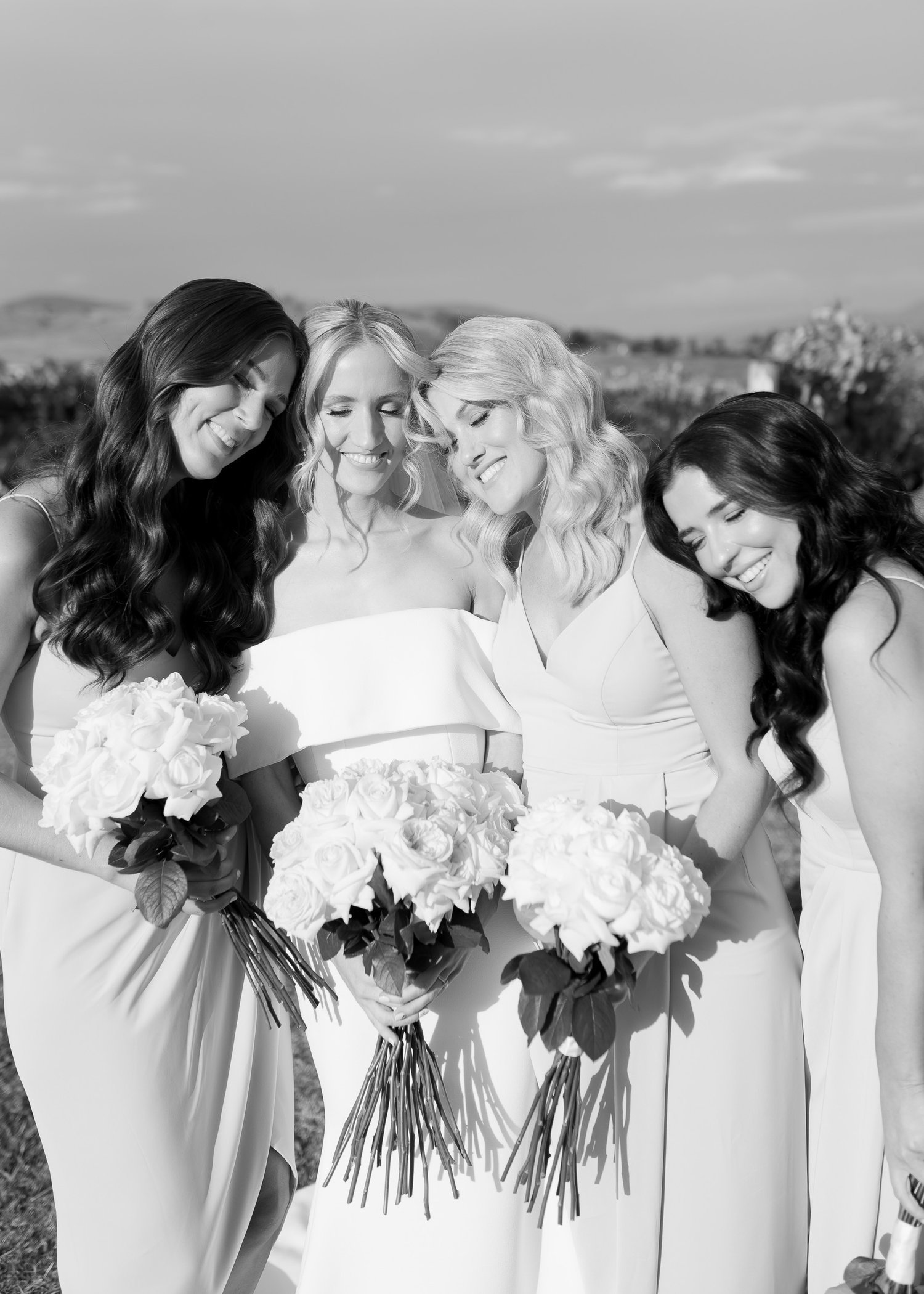 Four women in white dresses holding bouquets, standing closely together outdoors, smiling at the camera. Black and white photograph.