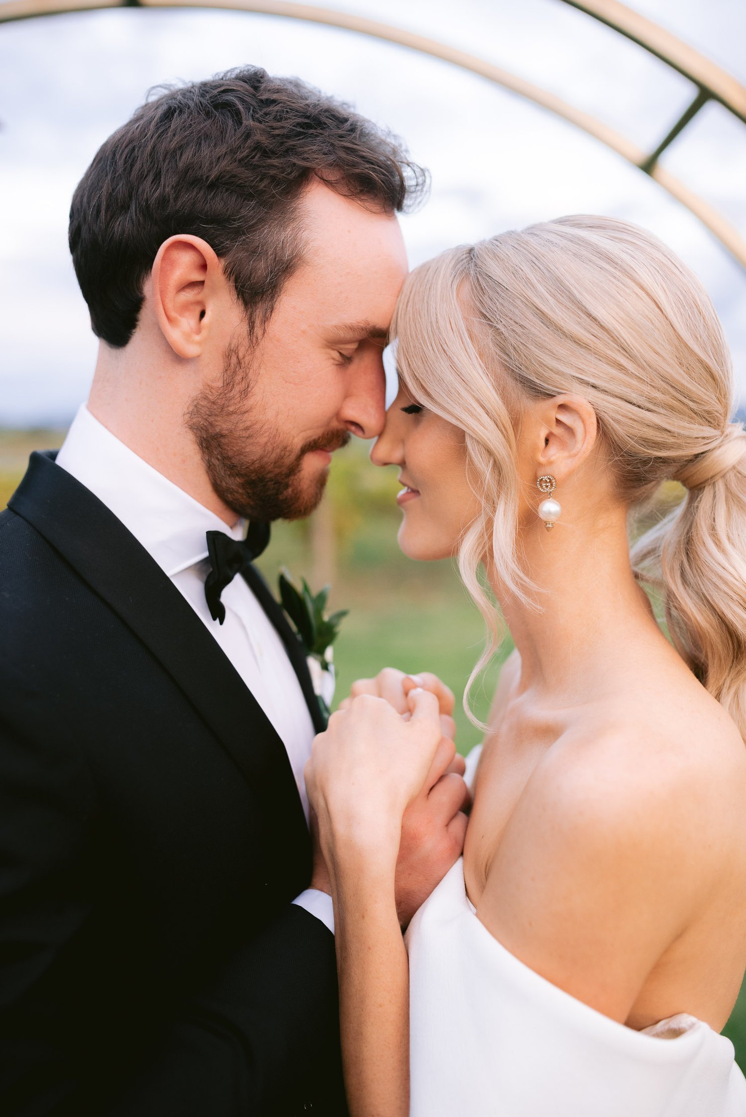 Close-up of a bride and groom leaning their foreheads together, smiling softly, with their hands clasped. The bride has blonde hair styled in a ponytail and wears a white off-the-shoulder dress, pearl earrings, and the groom is in a black suit with a