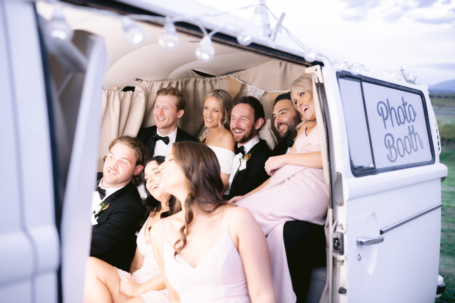 Group of people in a photo booth at an outdoor event, dressed in formal attire, smiling and laughing.