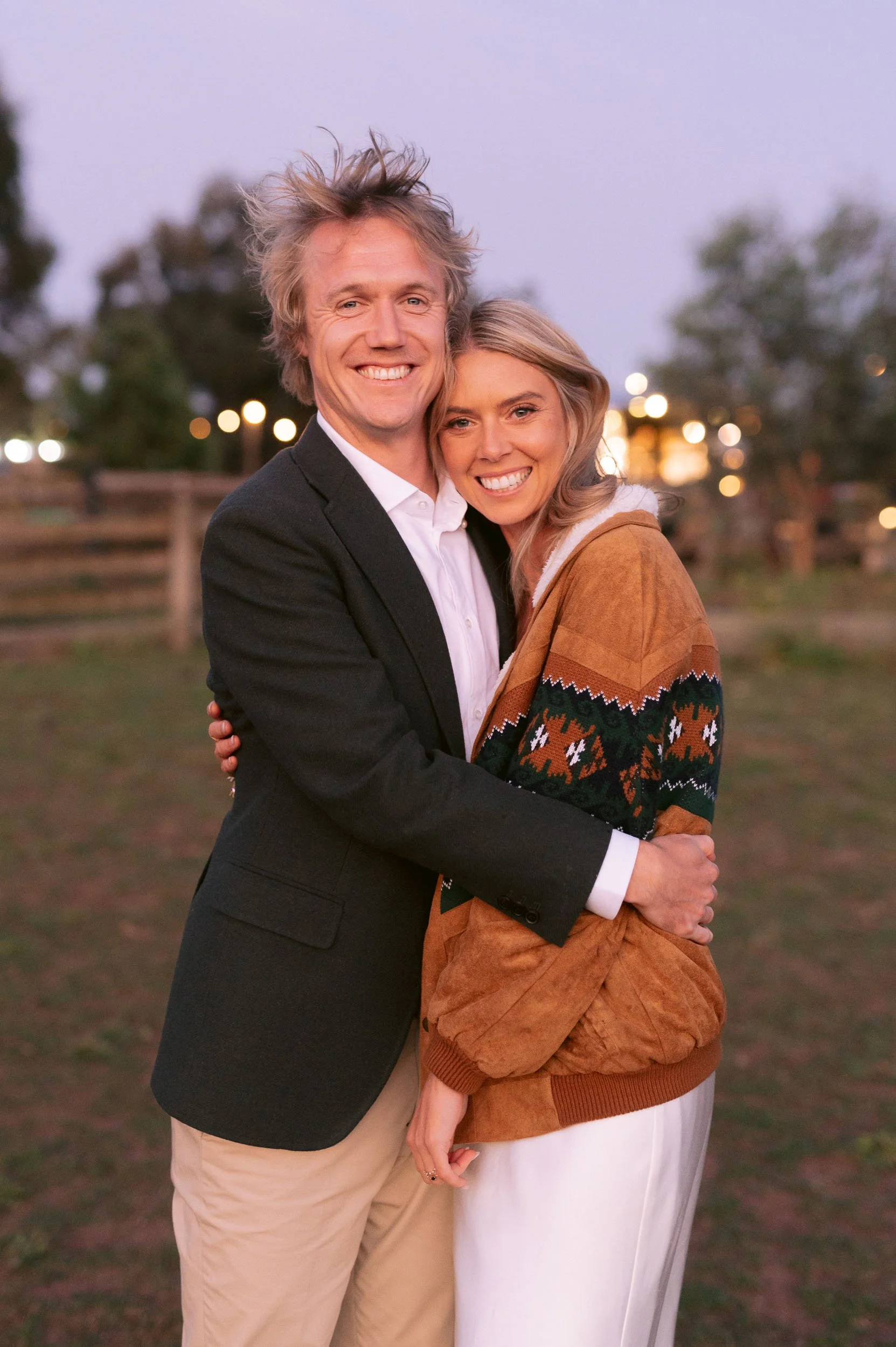 A happy couple hugging outdoors during sunset, with blurred trees and lights in the background at Quirindi Stables.