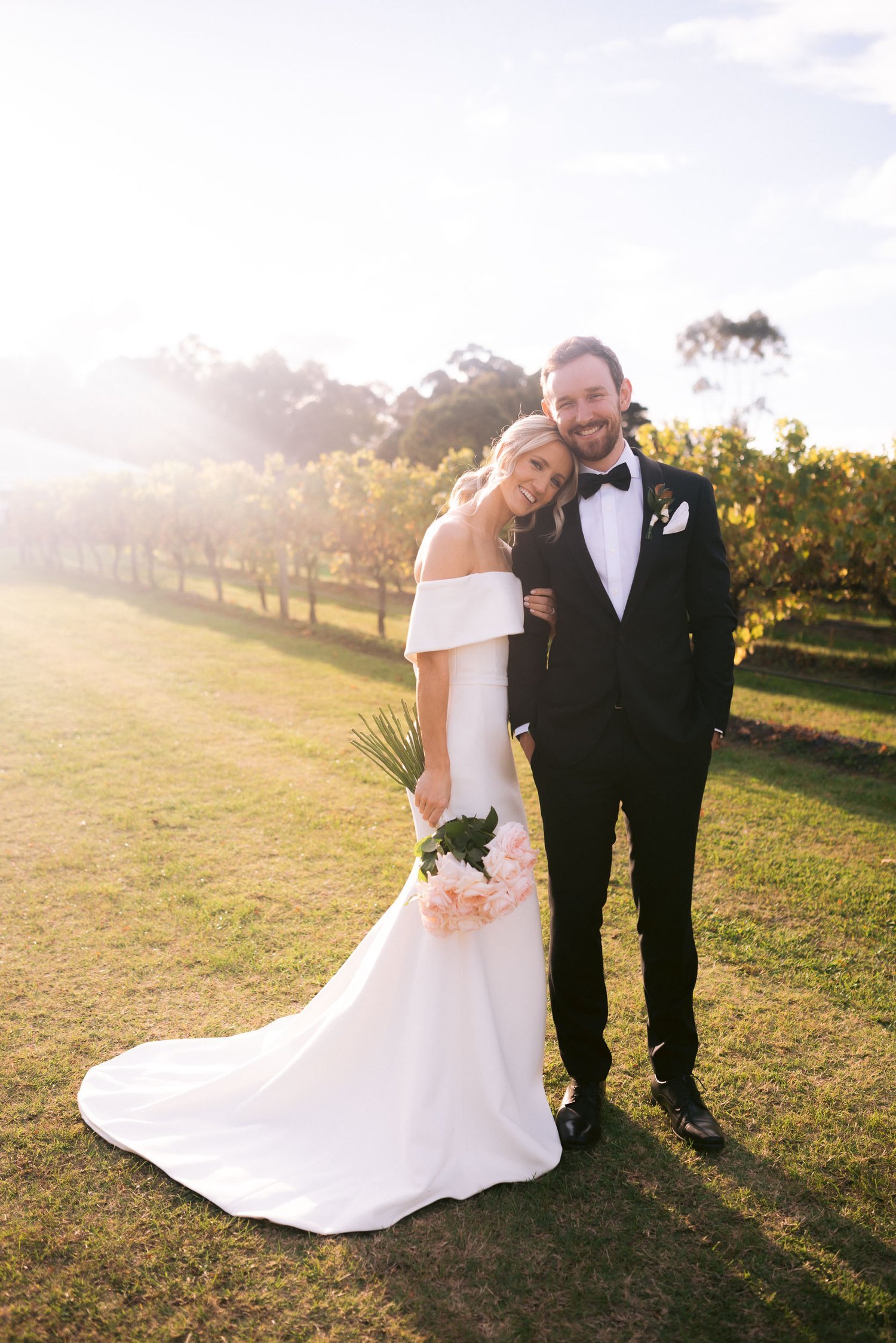 Bride and groom standing in a vineyard, bride in white dress holding flowers, groom in black suit, smiling, sunny day.