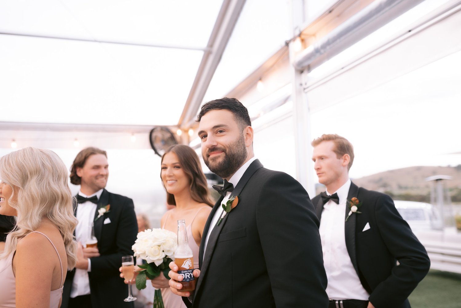 Group of elegantly dressed people at an outdoor event, with men in tuxedos and women in formal dresses, some holding drinks and a flower bouquet, under a tent with string lights.