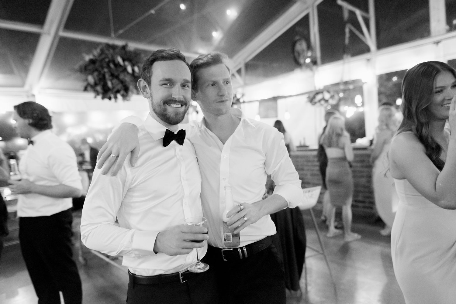 Two men in formal attire posing at a social event; one is wearing a bow tie and holding a wine glass, the other has an arm around him and holds a drink bottle. They are smiling in a festive setting.