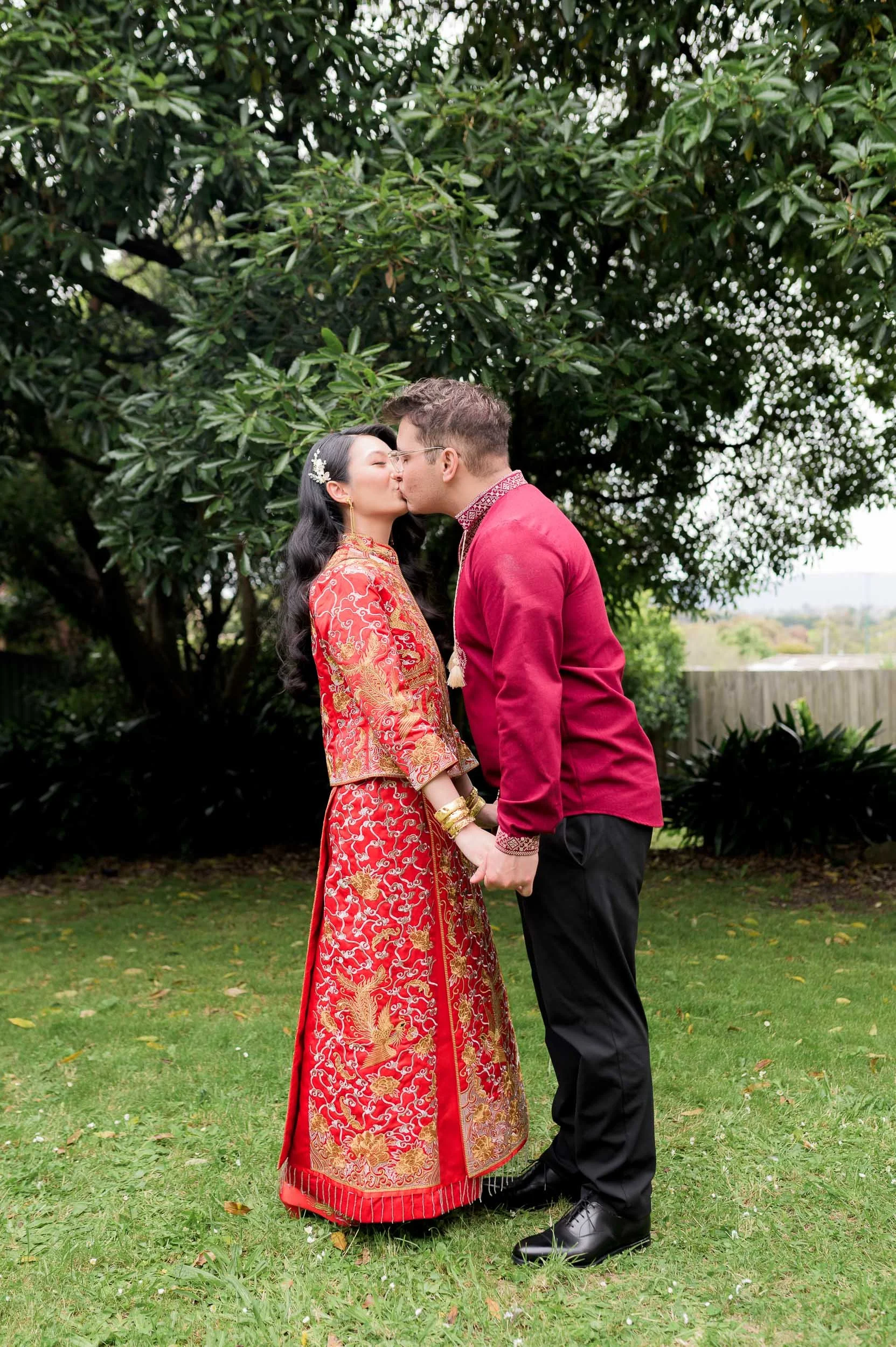 A couple in traditional red Chinese attire kissing in a garden.